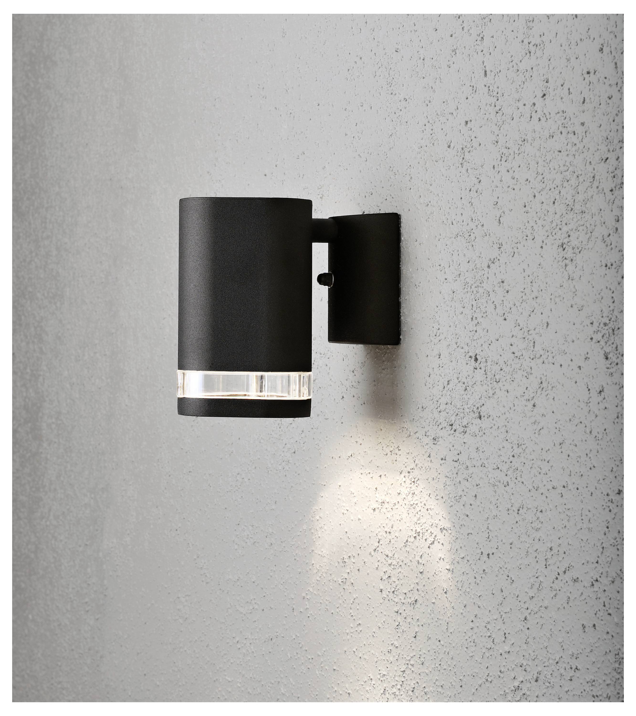 Eine moderne, zylindrische schwarze Wandleuchte mit klarem Glasunterteil, die Licht gegen eine strukturierte graue Wand abstrahlt.
