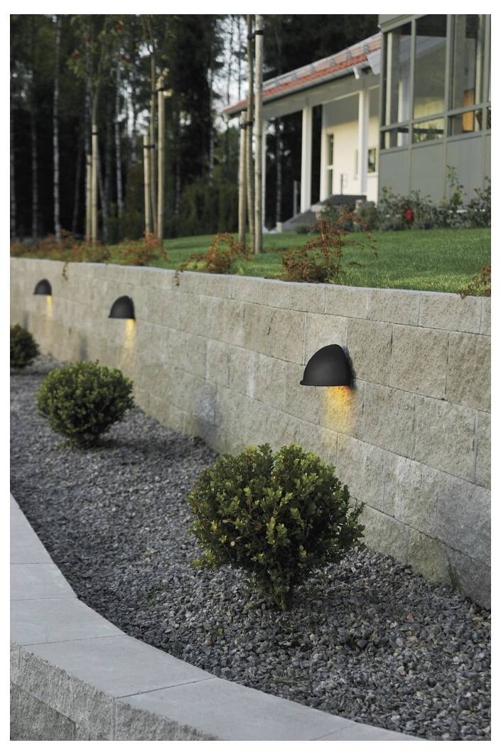 Ein modernes Haus mit einer gestalteten Gartenanlage, gekennzeichnet durch eine Natursteinmauer mit eingelassenen halbkreisförmigen Leuchten und sorgfältig geschnittenen Büschen.