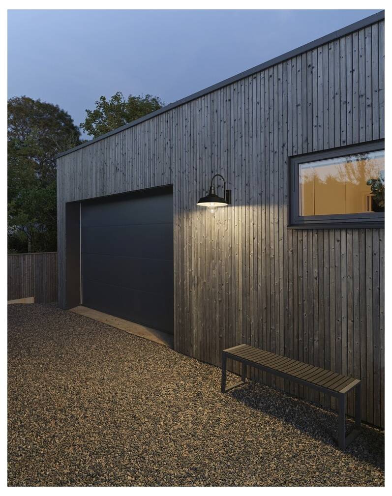 Ein modernes Holzgebäude mit einem großen Garagentor, einer Außenlampe und einer Bank auf einer Kiesfläche, umgeben von Bäumen bei Einbruch der Dämmerung.