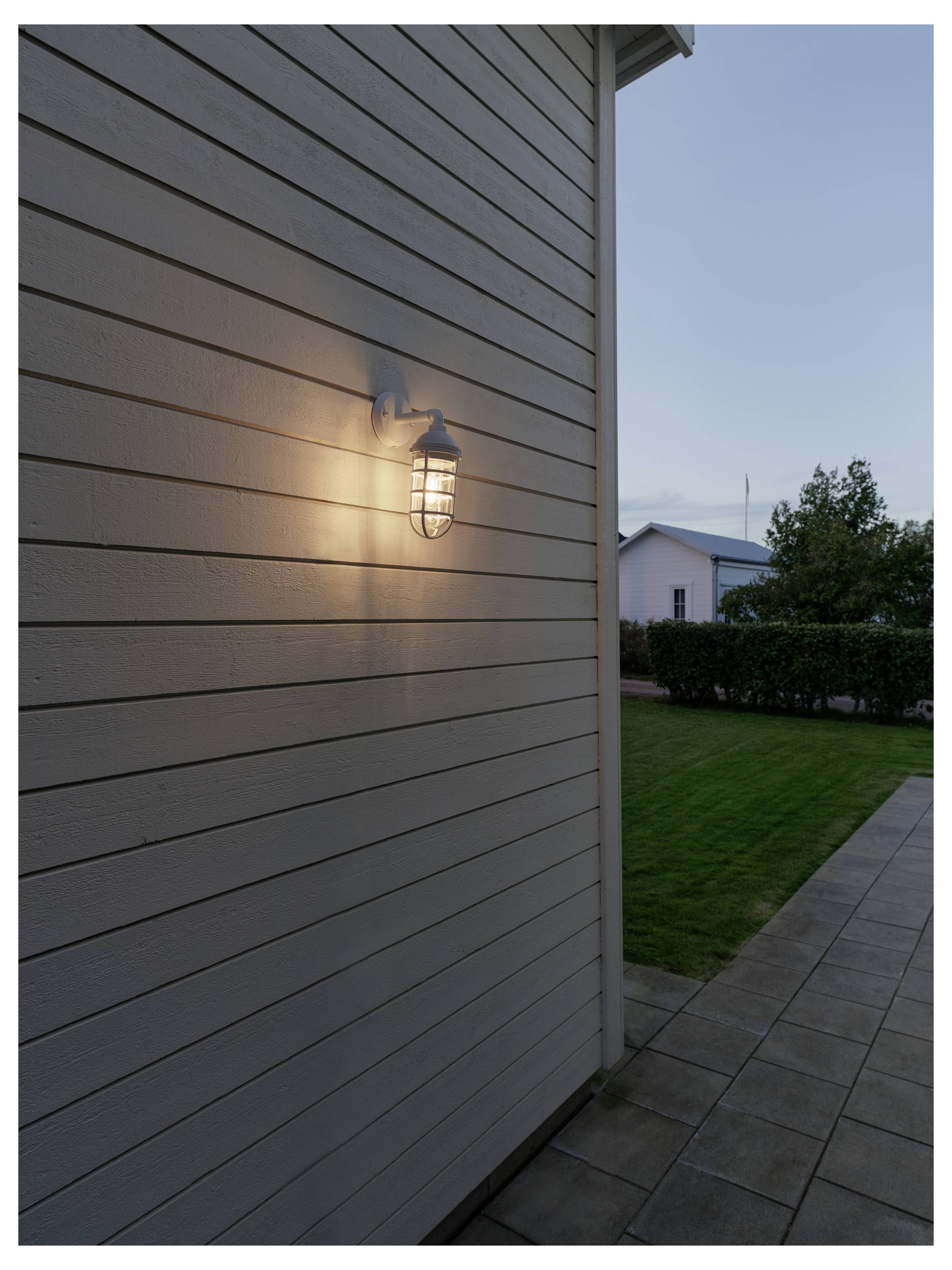 Eine leuchtende Wandlampe an der Hausfassade beleuchtet eine Holzvertäfelung in der Abenddämmerung, mit einer Rasenfläche und einer Terrasse im Hintergrund.