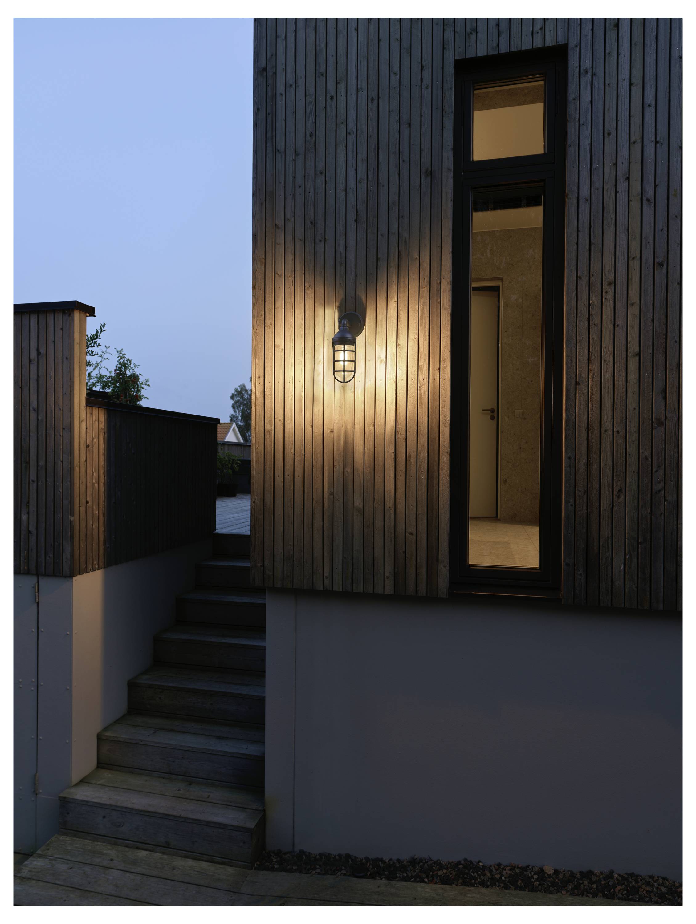 Außenansicht eines modernen Hauses in der Abenddämmerung, mit einem beleuchteten Fenster und einer Wandleuchte, die eine Holzvertäfelung und eine Treppe nach oben beleuchtet.