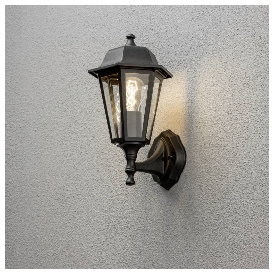 Eine schwarze Metall-Wandlaterne im Freien strahlt ein warmes Licht vor einer schlichten grauen Wand.