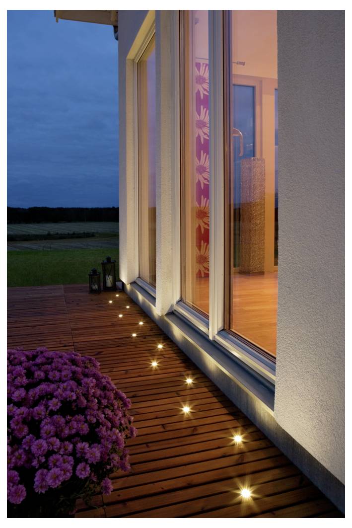 Eine moderne Hausaußenansicht in der Abenddämmerung mit großen Glasfenstern, die ein warmes Innenlicht ausstrahlen. Die Outdoor-Terrasse ist mit kleinen Bodenleuchten und Kübelpflanzen dekoriert.