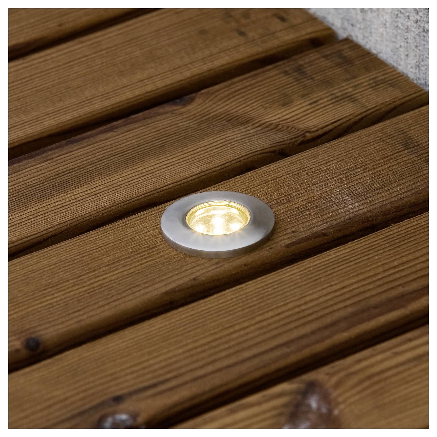 Eine runde, eingelassene LED-Leuchte, die in einer Holzdielen-Planke eingebettet ist und den umgebenden Bereich beleuchtet, wahrscheinlich für den Außeneinsatz oder zur Schaffung einer Atmosphäre.