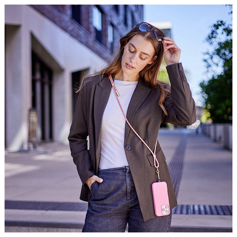 Eine Person steht außerhalb eines Gebäudes, trägt eine Jacke und Jeans, mit einem Smartphone an einem rosa Riemen um den Hals.