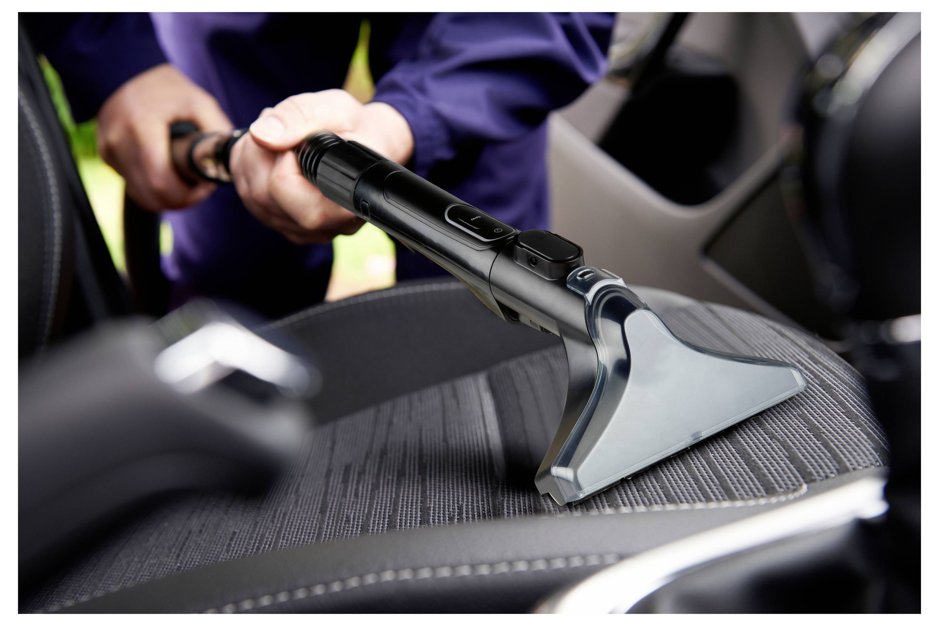 Eine Person verwendet ein Staubsauger-Zubehör, um den Stoffsitz eines Autos zu reinigen und konzentriert sich dabei darauf, Schmutz und Staub von Innenoberflächen zu entfernen.