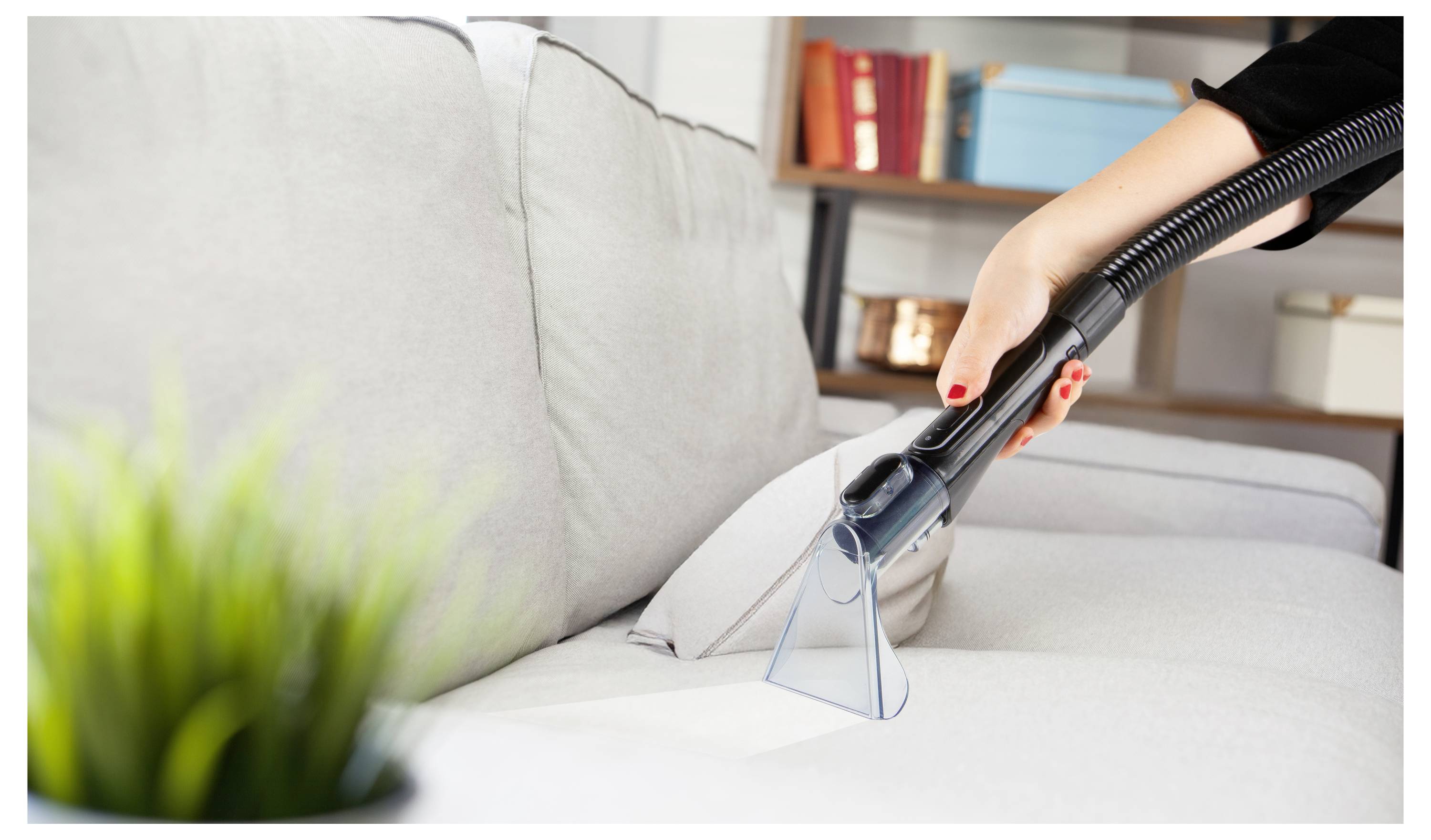 Eine Hand benutzt ein Saugrohraufsatz, um ein graues Sofa zu reinigen, wobei ein Teil eines Bücherregals im Hintergrund zu sehen ist.