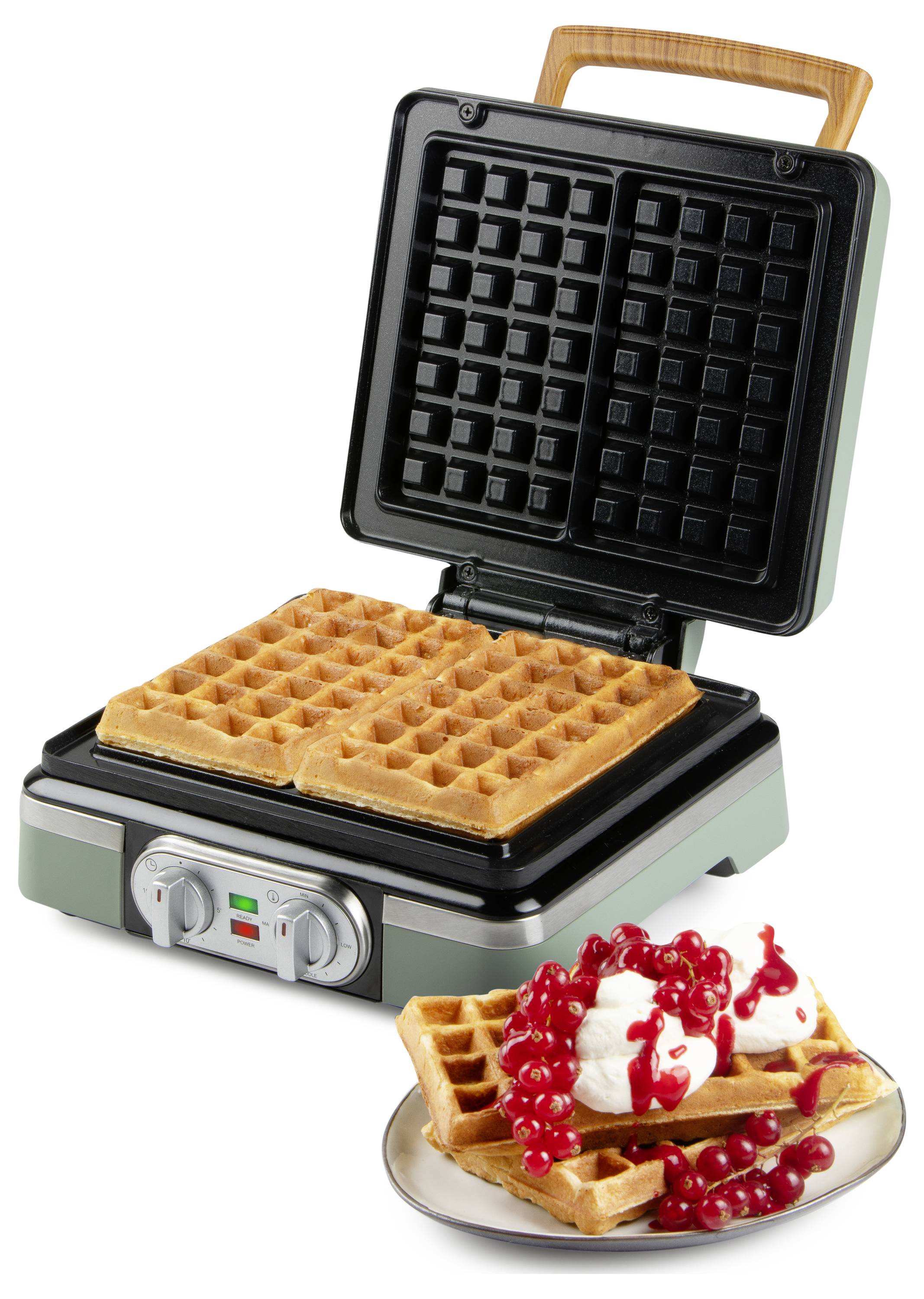 Ein Waffeleisen mit zwei fertigen Waffeln im Inneren. Ein Teller mit Waffeln, die mit Schlagsahne und roten Beeren belegt sind, befindet sich im Vordergrund.
