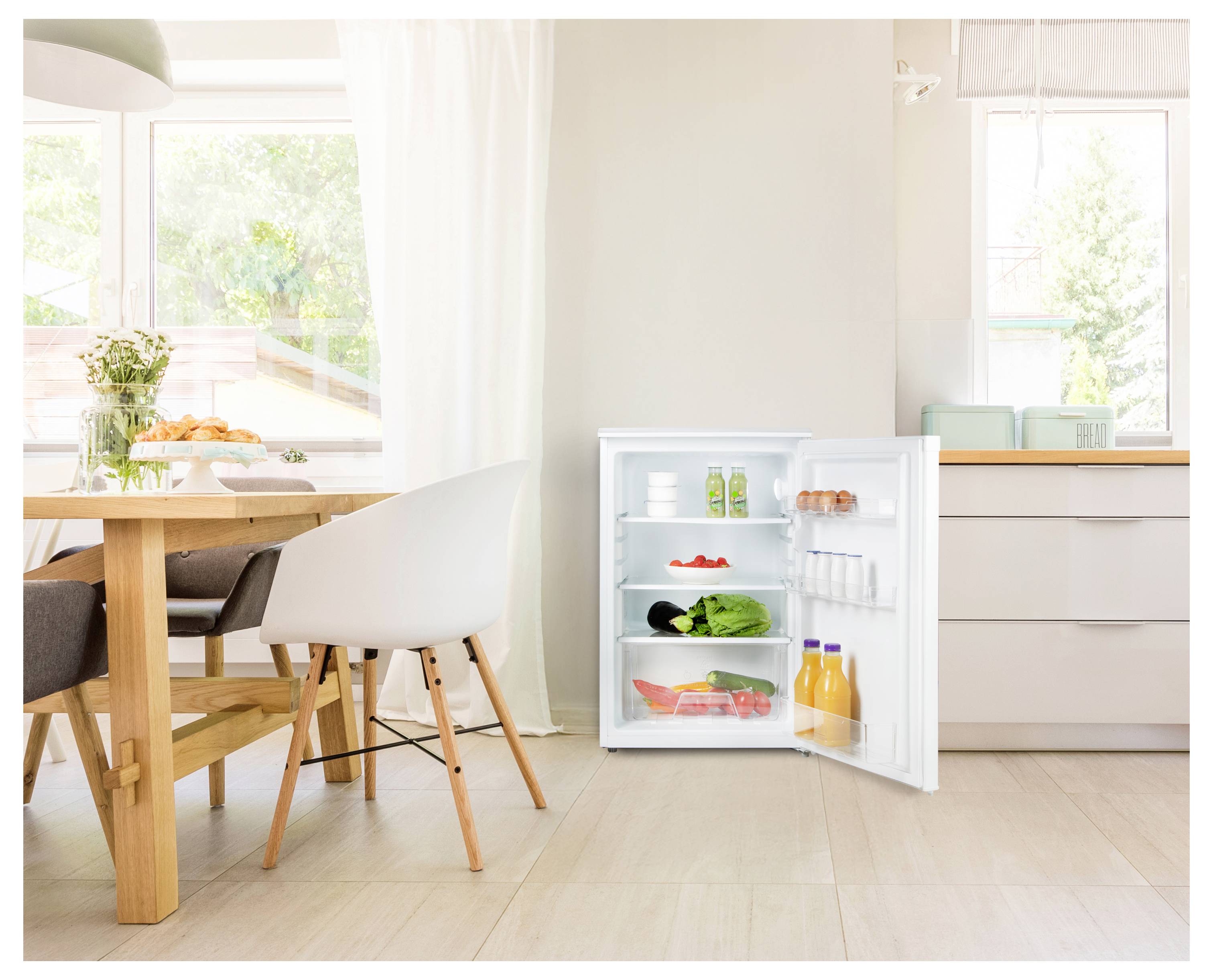 Ein offener kleiner Kühlschrank in einer hellen Küche mit Getränken und Gemüse im Inneren, neben einem Esstisch, der an einem großen Fenster steht.