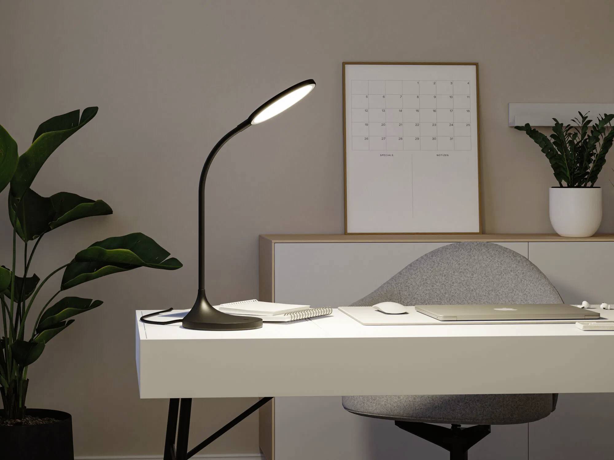 Eine elegante moderne Schreibtischlampe beleuchtet einen weißen Schreibtisch mit einem Laptop, einem Notizblock und einer Topfpflanze. An der Wand hängt ein Kalender.
