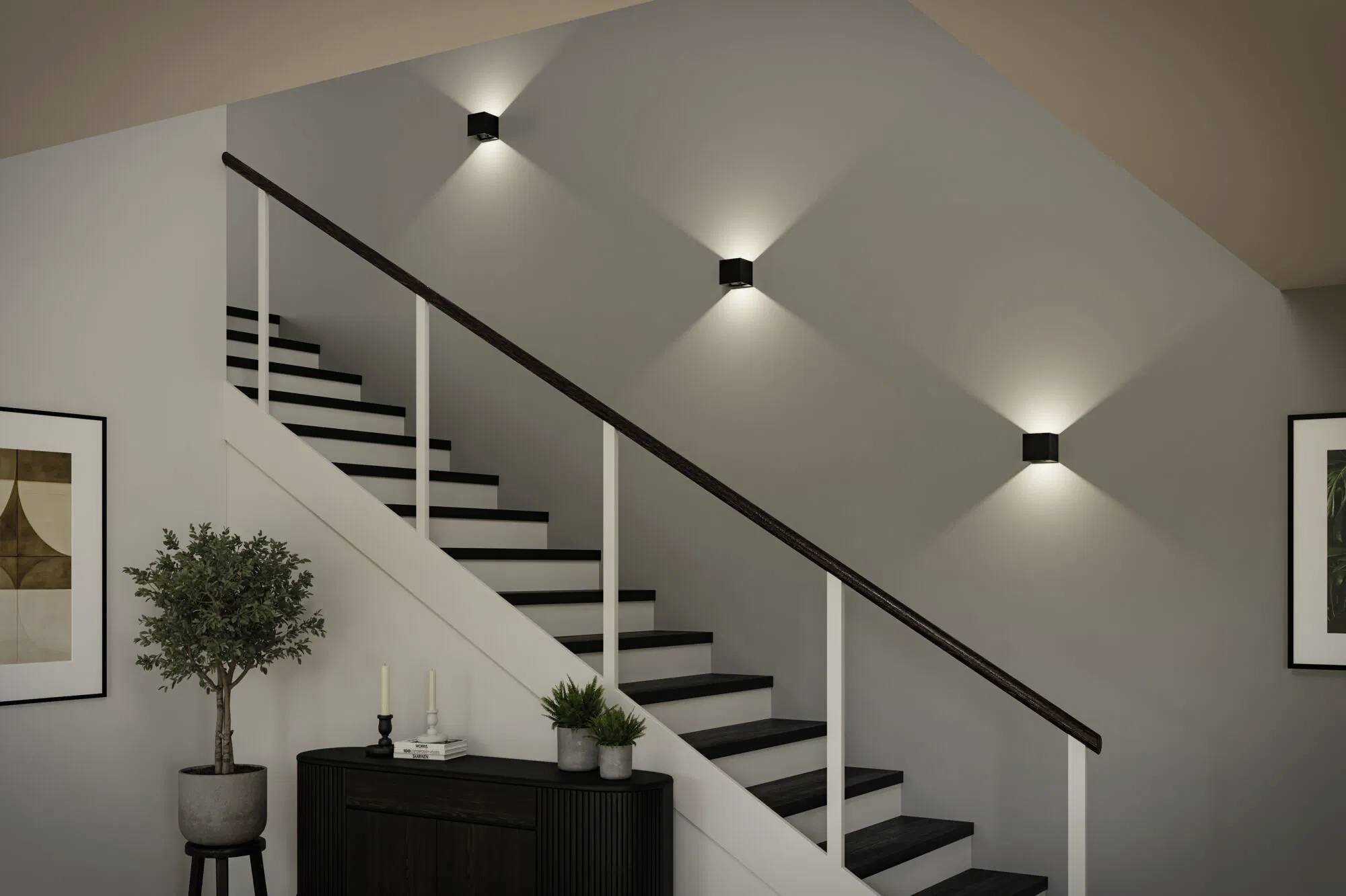 Eine moderne Treppe mit schwarzen und weißen Stufen, beleuchtet von an der Wand montierten Leuchten. Ein kleiner Schrank und eine Pflanze befinden sich am Fuß der Treppe.