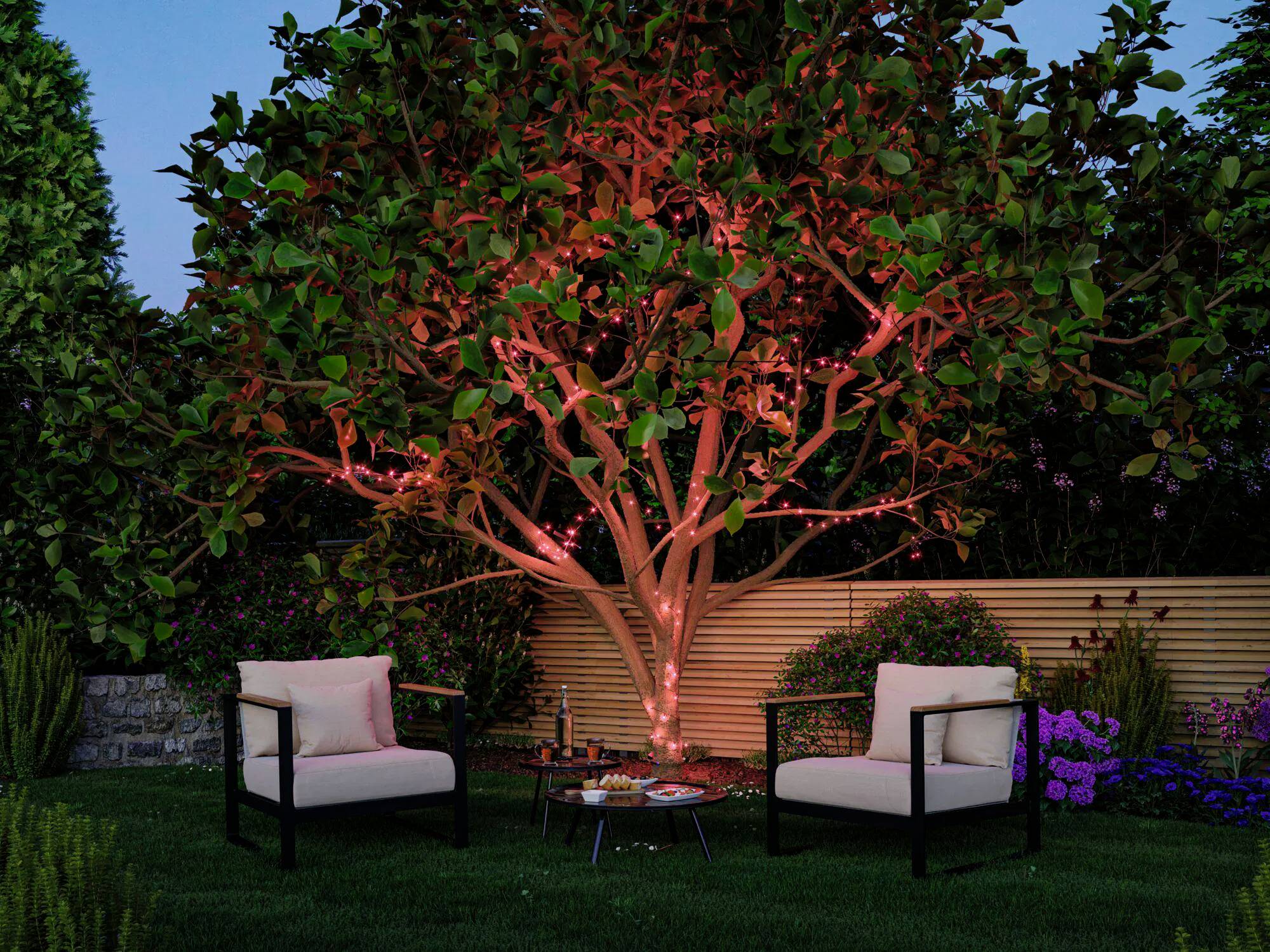 Eine Gartenszene mit zwei gepolsterten Stühlen auf einem Rasen, einem dekorierten Tisch mit Snacks und Weingläsern und einem Baum, der mit roten Lichtern beleuchtet ist.
