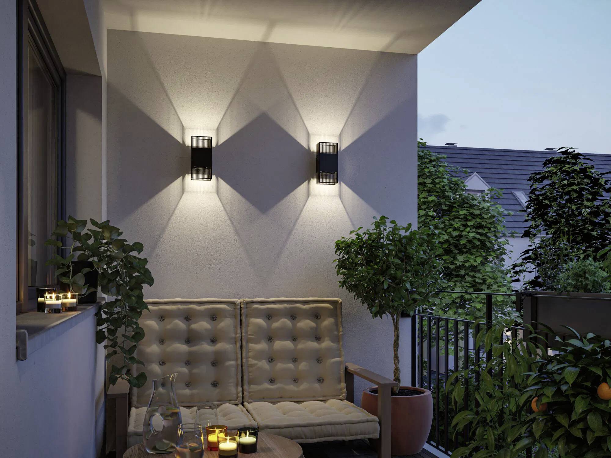 Eine gemütliche Balkonszenerie in der Abenddämmerung mit zwei beleuchteten Wandleuchten, die Lichtmuster werfen, einem gepolsterten Sofa, einem kleinen Tisch und Topfpflanzen.