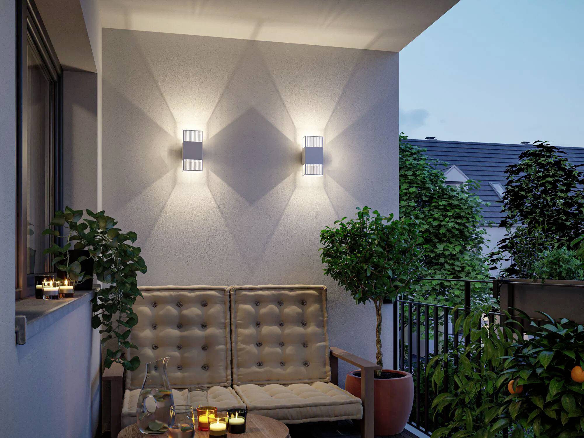 Eine gemütliche Terrasse im Freien bei Einbruch der Dämmerung mit zwei Wandleuchten, die geometrische Schatten werfen. Ein gepolsterter Sitzplatz, ein kleiner Tisch mit Kerzen und Topfpflanzen.