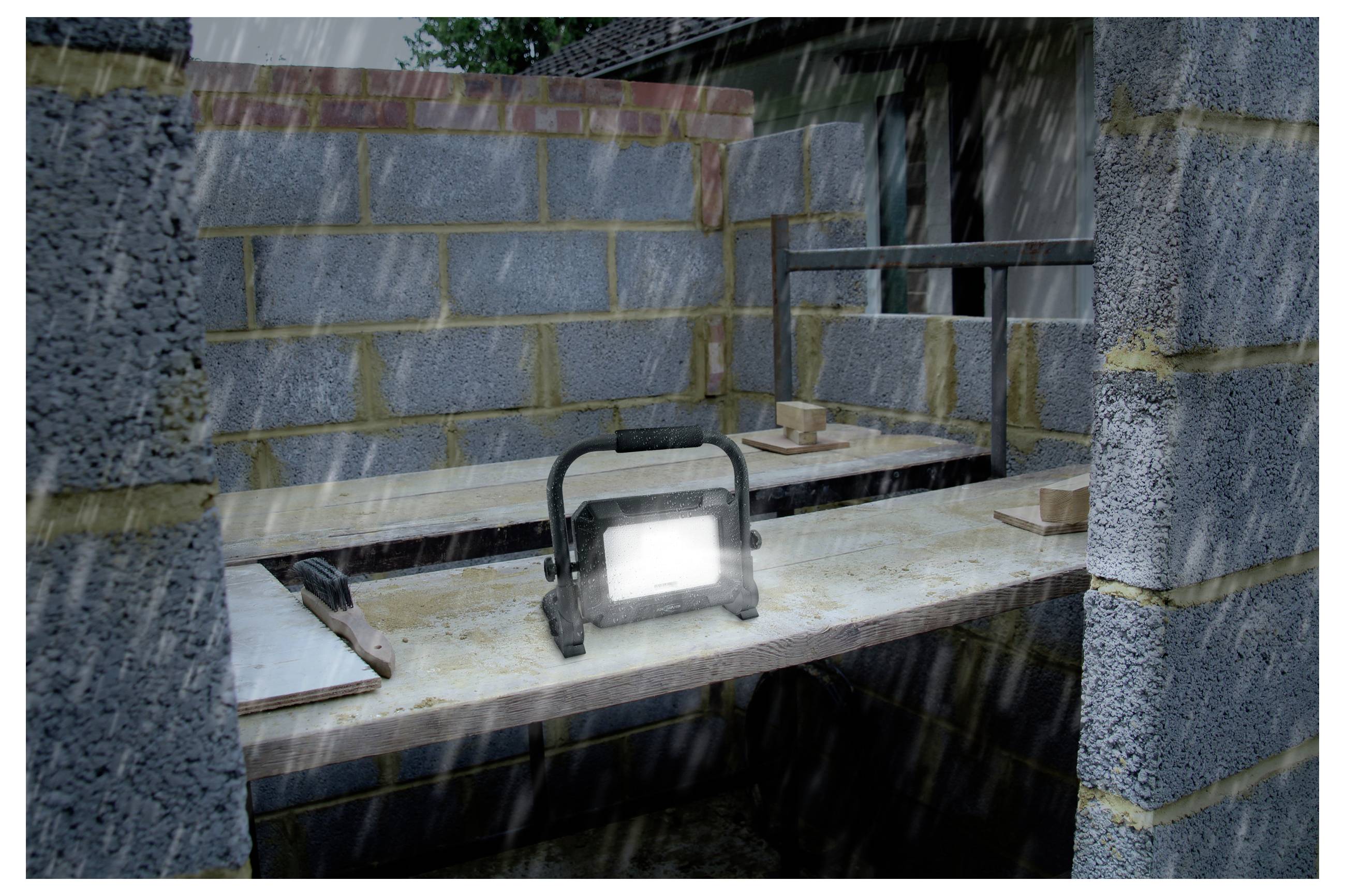 Eine Baustelle mit teilweise errichteten Wänden und einer Werkbank unter Regen. Ein helles, tragbares Arbeitsscheinwerfer beleuchtet die Szenerie.