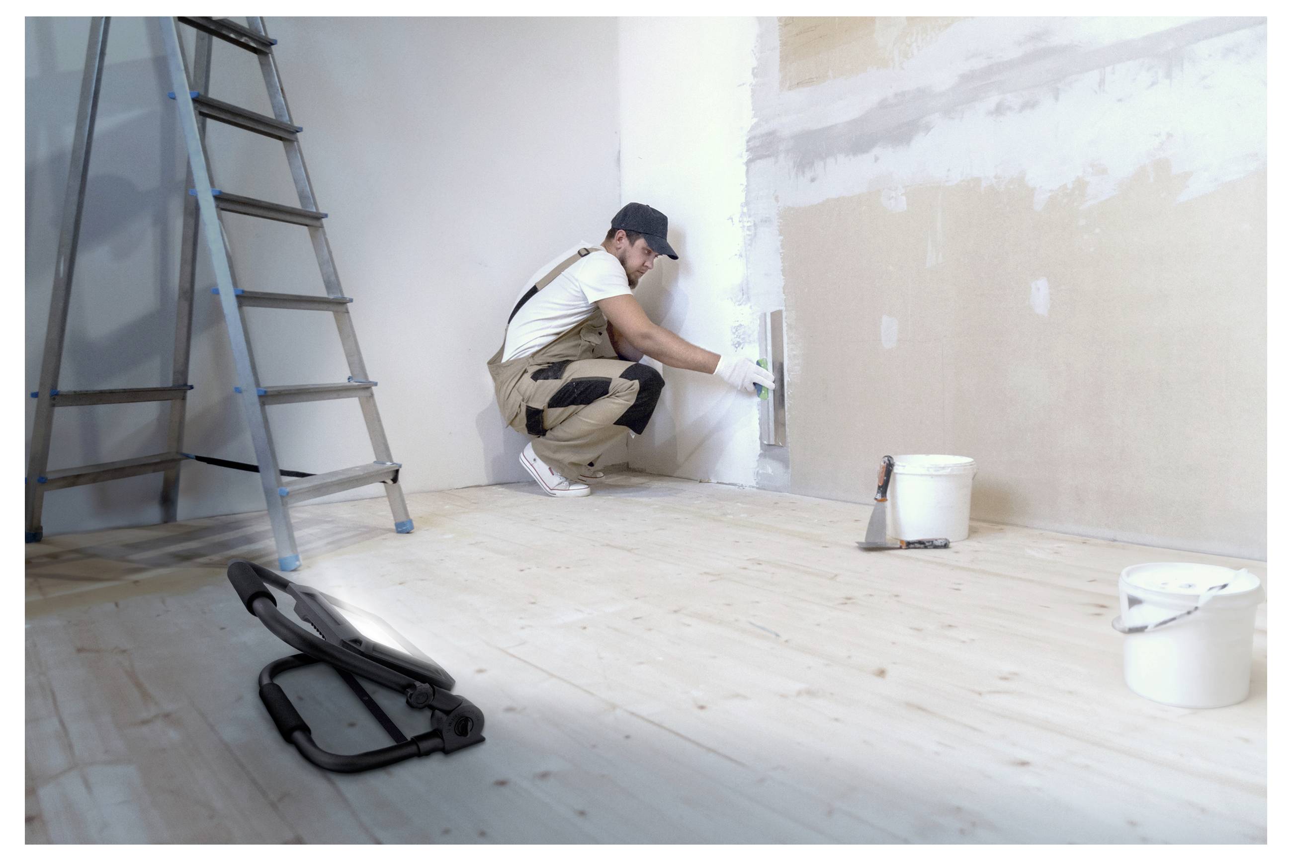 Eine Person, die Putz an eine Wand in einem Raum mit Holzboden aufträgt. Eine Leiter und Farbkübel befinden sich in der Nähe und deuten auf eine Renovierung hin.