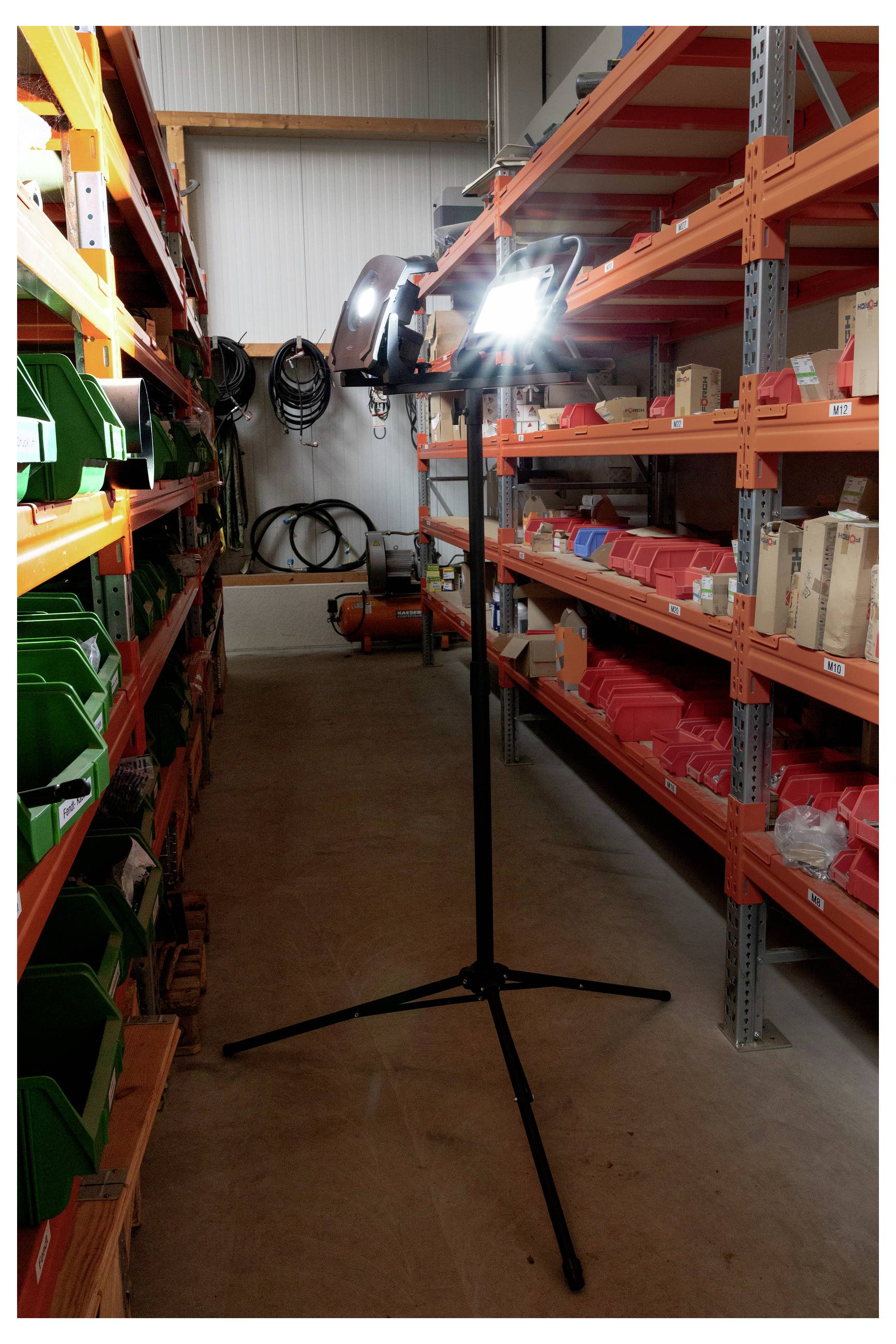 Ein gut beleuchteter Lagerhallengang mit orangefarbenen Metallregalen auf beiden Seiten, die verschiedene Behälter und Gegenstände tragen; eine helle Lampe steht in der Mitte.