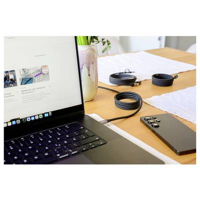 Ein Laptop auf einem Tisch, der über ein Kabel mit einem Smartphone verbunden ist, mit aufgewickelten Kabeln und einer Vase mit grünen Blättern im Hintergrund.