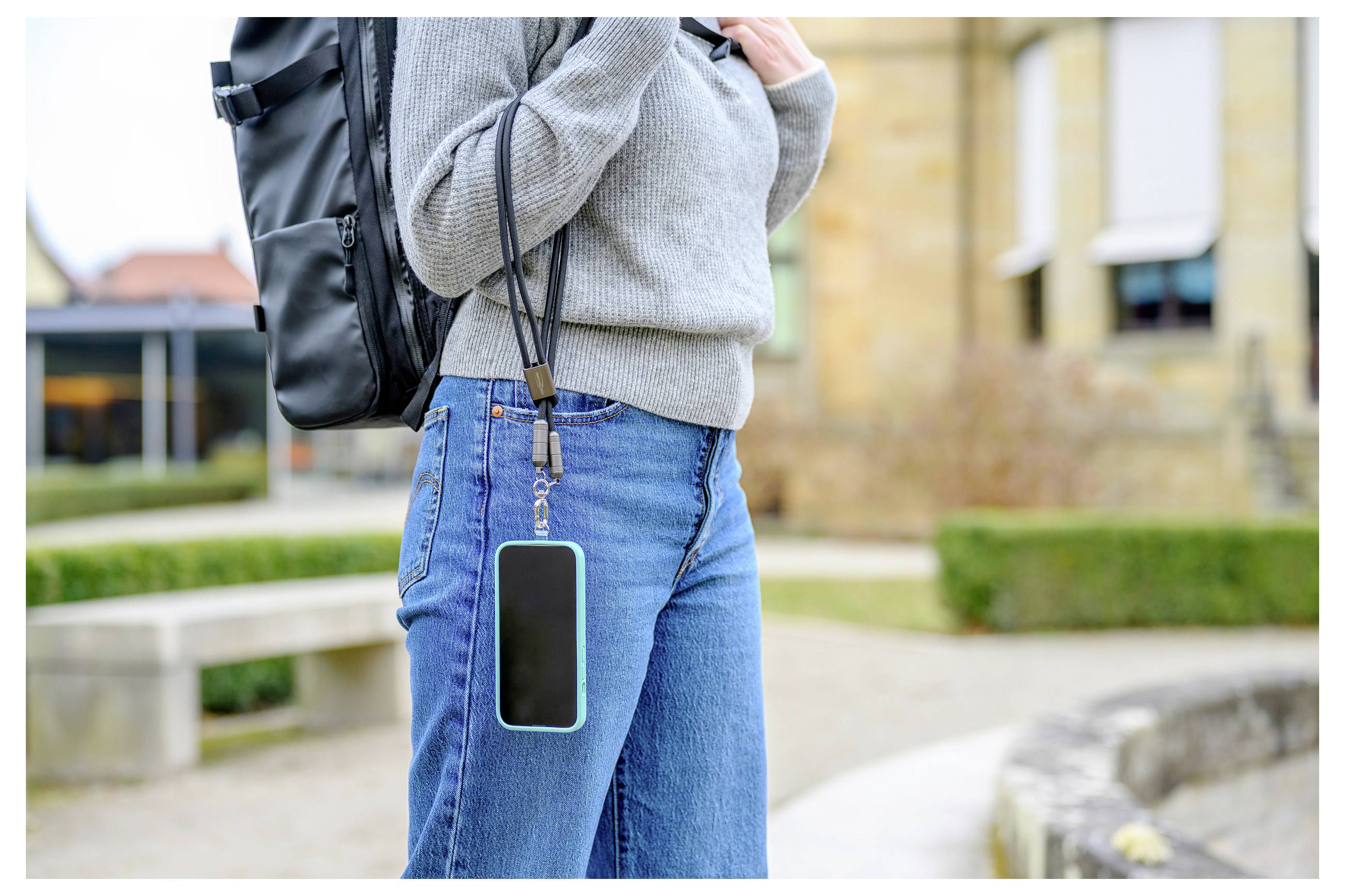 Person mit einem Rucksack und grauem Pullover im Freien, der ein Smartphone an einer Schnur um den Hals hält. Gebäude im Hintergrund.