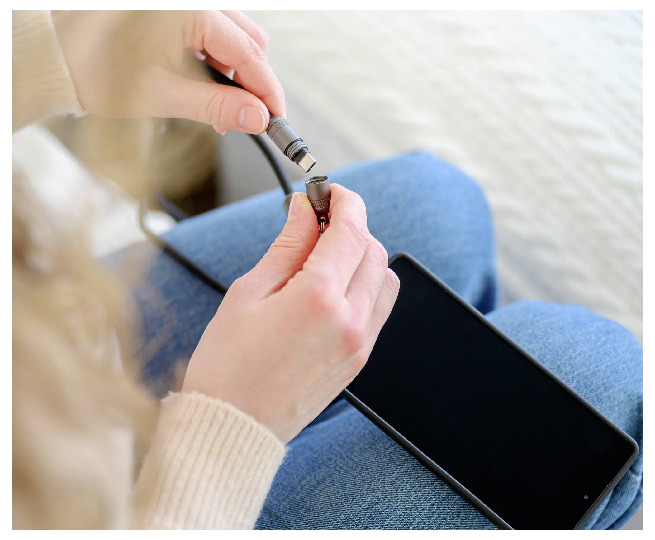 Eine Person steckt ein Kabel in ein Smartphone, wobei das Aufladen oder die Verbindung des Geräts hervorgehoben wird, während das Telefon auf ihrem mit Jeansstoff bekleideten Schoß liegt.