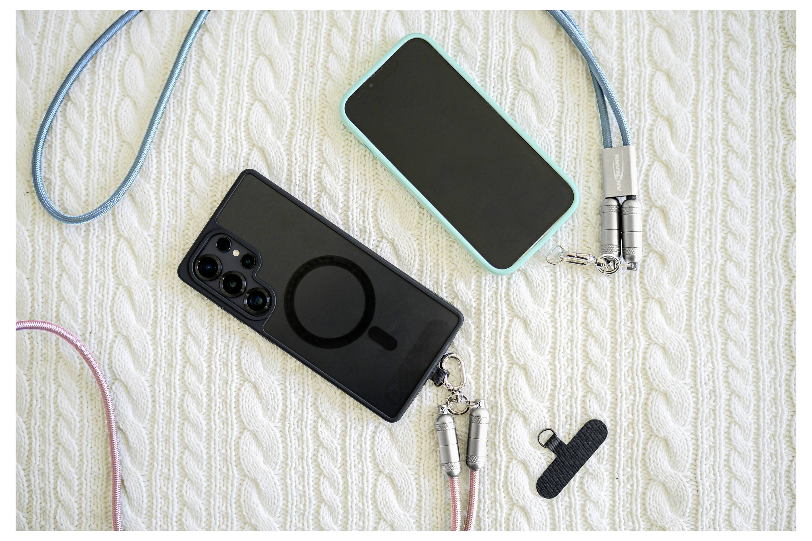 Zwei Smartphones auf einer strukturierten Oberfläche, jeweils mit farbigen Trageband befestigt. Ein Adapter liegt in der Nähe und deutet auf Laden oder Konnektivität hin.