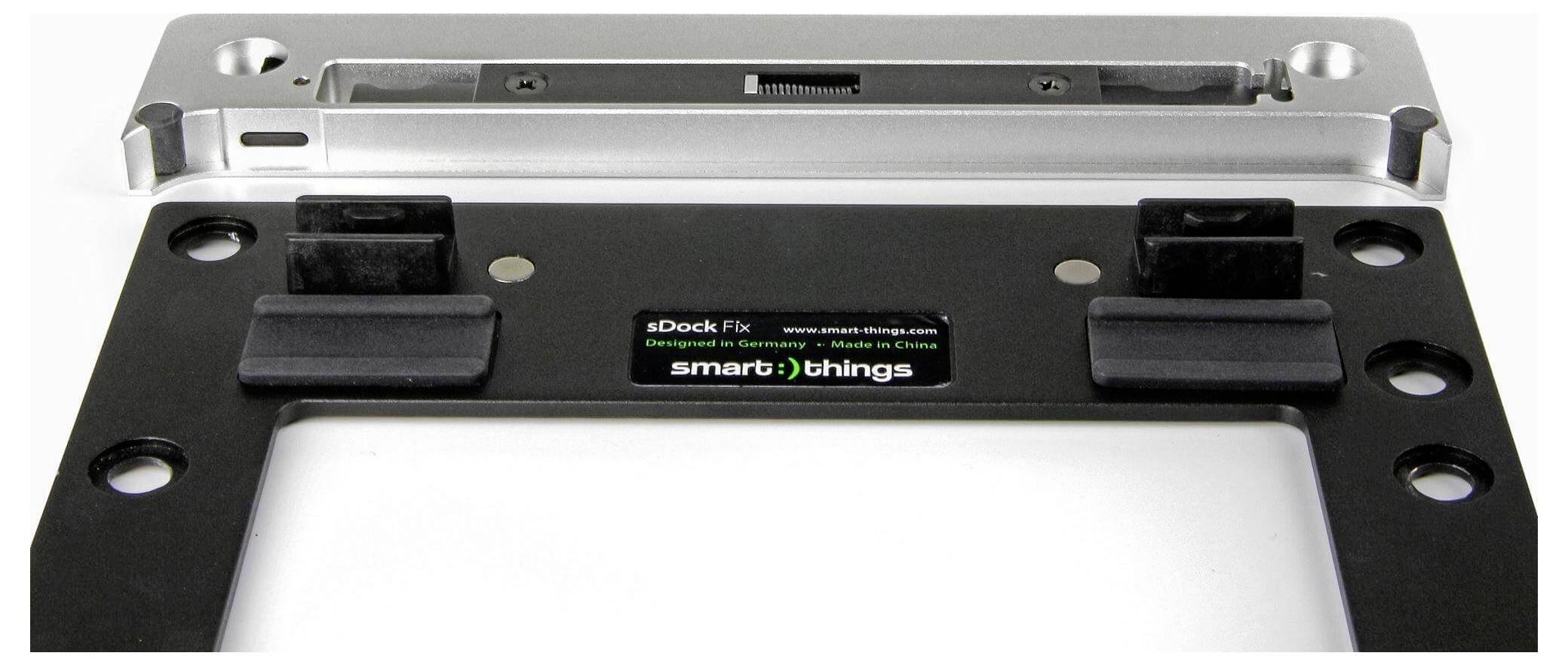 Eine silberne Metall-Laptop-Dockingstation mit einem schwarzen Sockel und zwei Befestigungshalterungen. Der Text lautet: 'sDock Fix, designed by smart:)things, Made in China.'