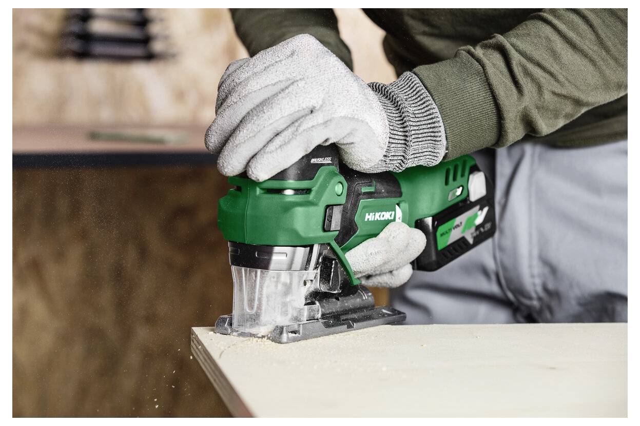 Eine Person verwendet eine grüne Stichsäge, um ein Holzbrett in einer Werkstattumgebung zu durchtrennen und trägt dabei Schutzhandschuhe.