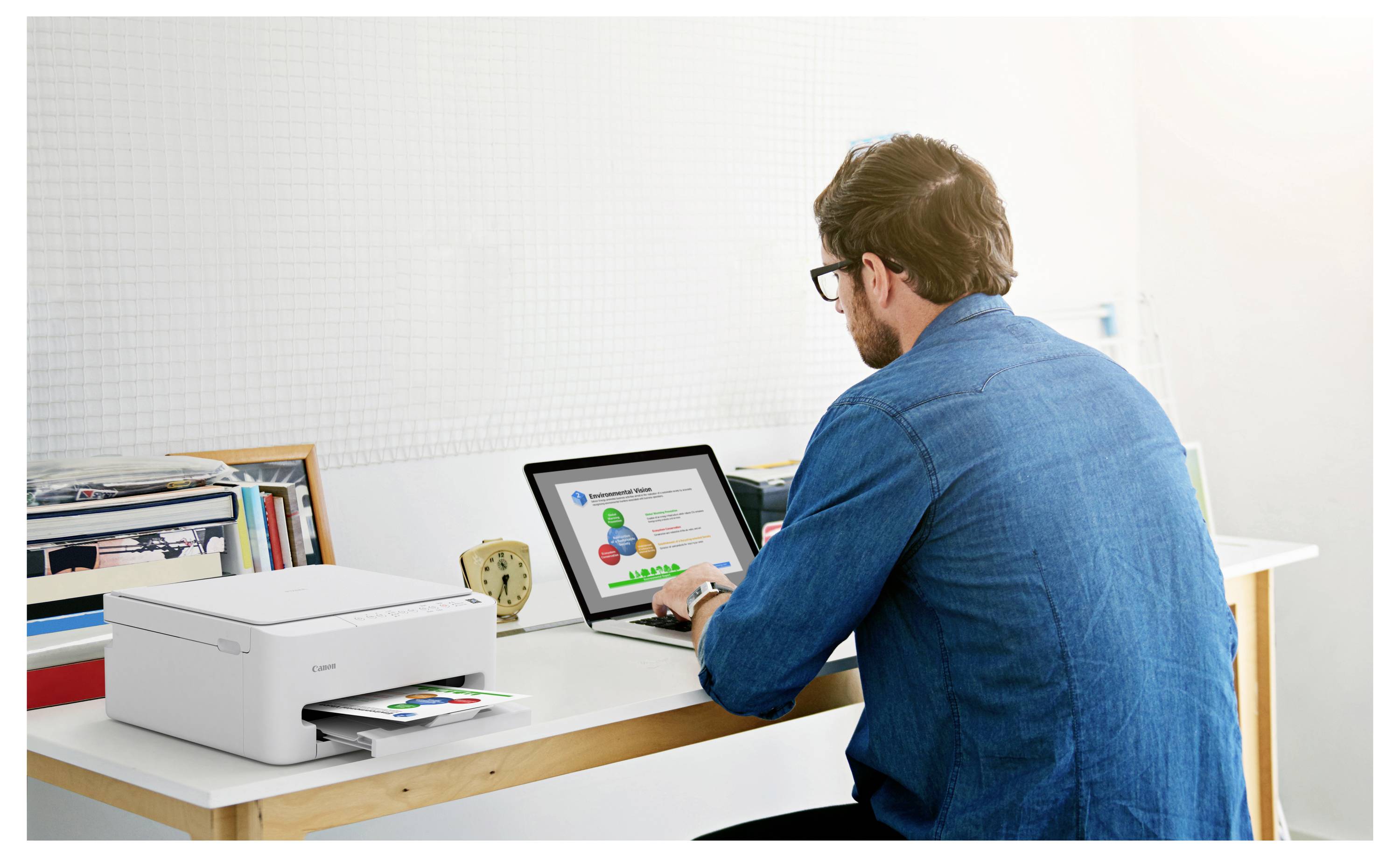 Eine Person sitzt an einem Schreibtisch und verwendet einen Laptop, neben dem ein Drucker steht. Auf dem Laptop-Bildschirm wird ein buntes Diagramm oder eine Infografik angezeigt.