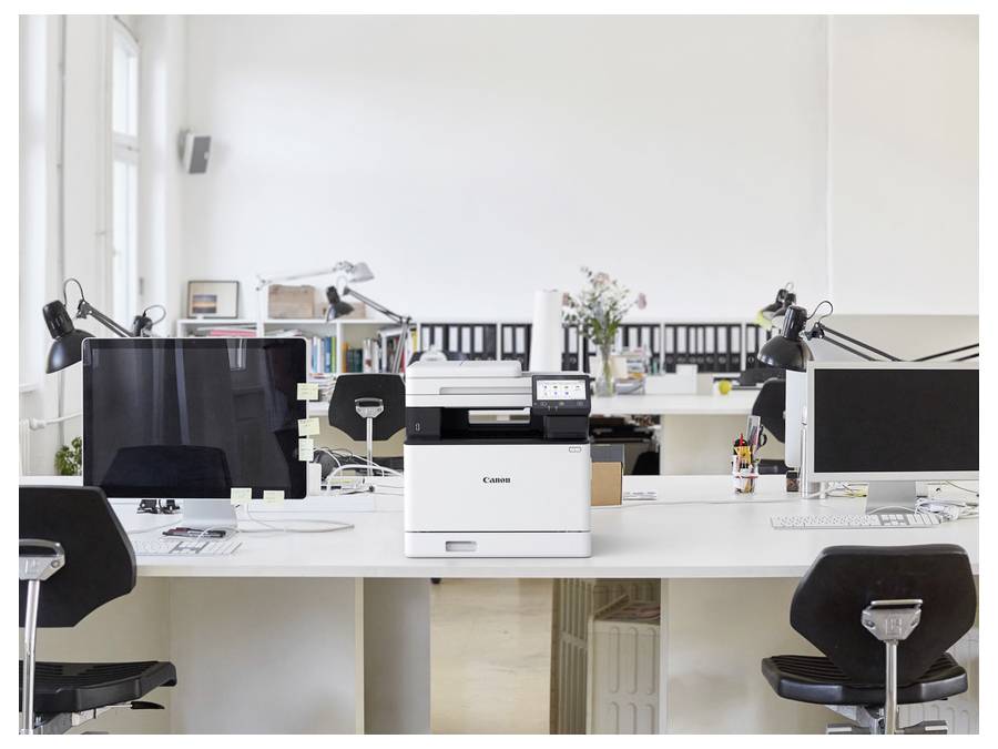 Modernes Büro mit Schreibtischen, Computern, Lampen und einem Drucker, der auf einem Tisch in einem hellen, offenen Raum mit Büchern und Accessoires platziert ist.