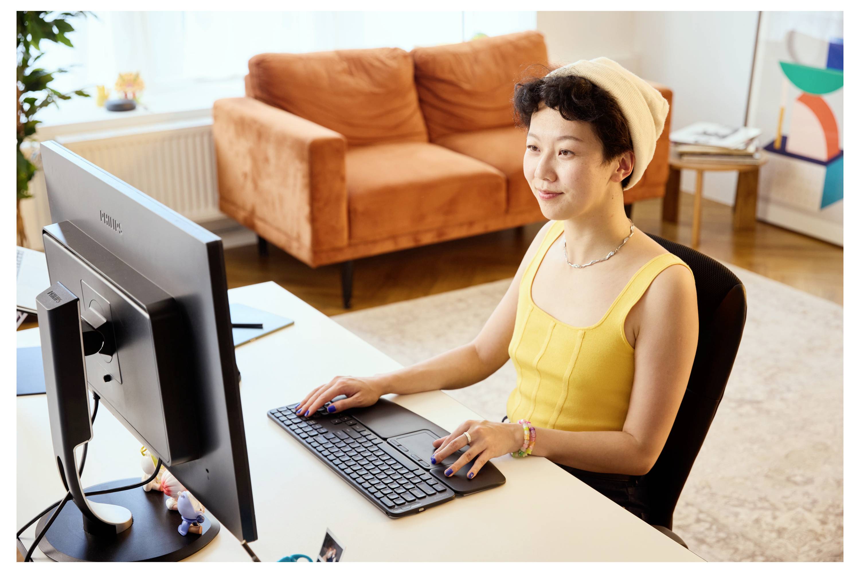 Eine Person sitzt an einem Schreibtisch und lächelt, während sie an einem Computer arbeitet. Sie befindet sich in einem hellen, modernen Raum mit einem pfirsichfarbenen Sofa hinter sich.