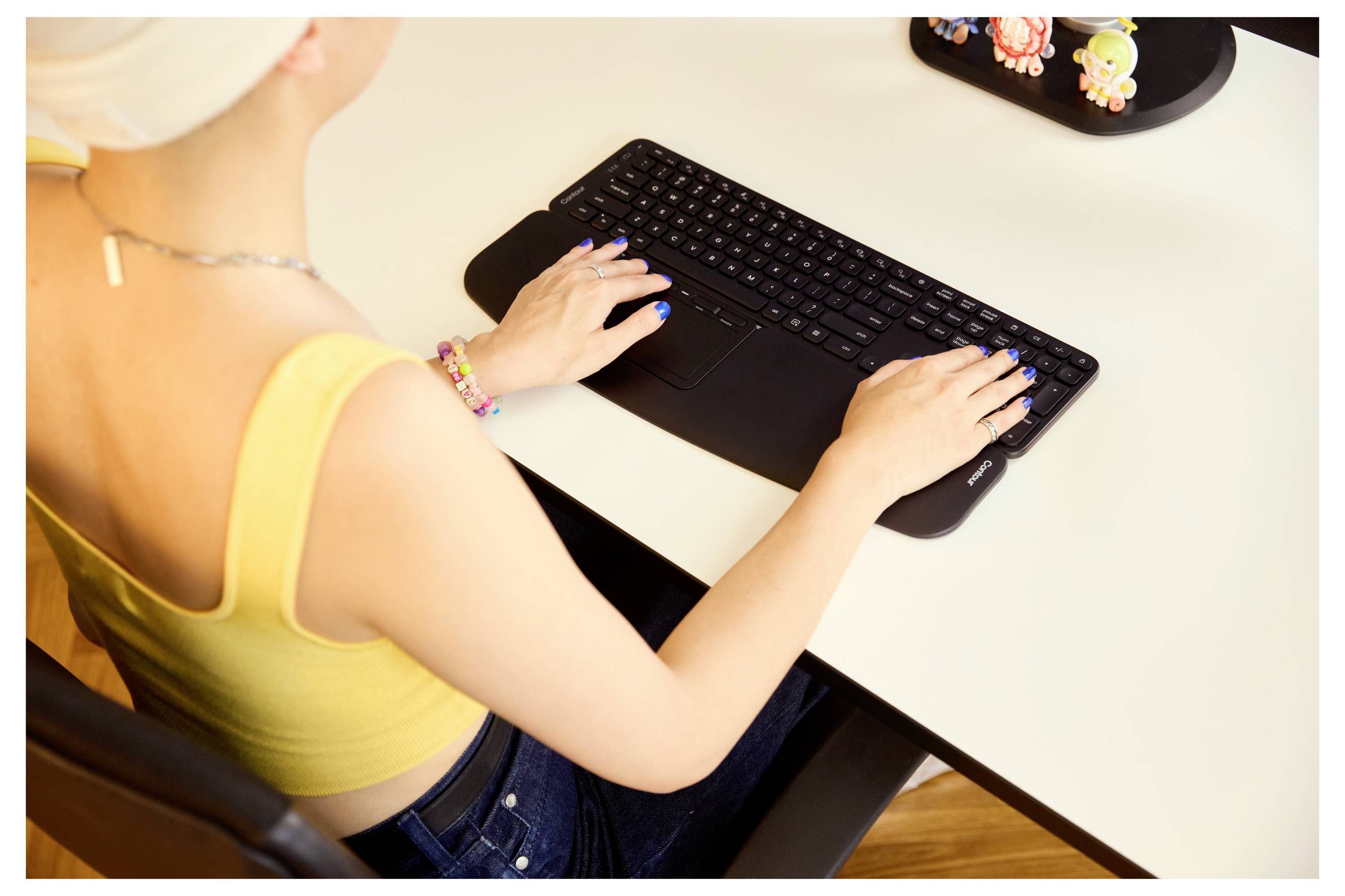 Eine Person in einem gelben Oberteil sitzt an einem Schreibtisch und tippt auf einer schwarzen Tastatur. Bunte Accessoires sind an den Händen und auf dem Schreibtisch sichtbar.