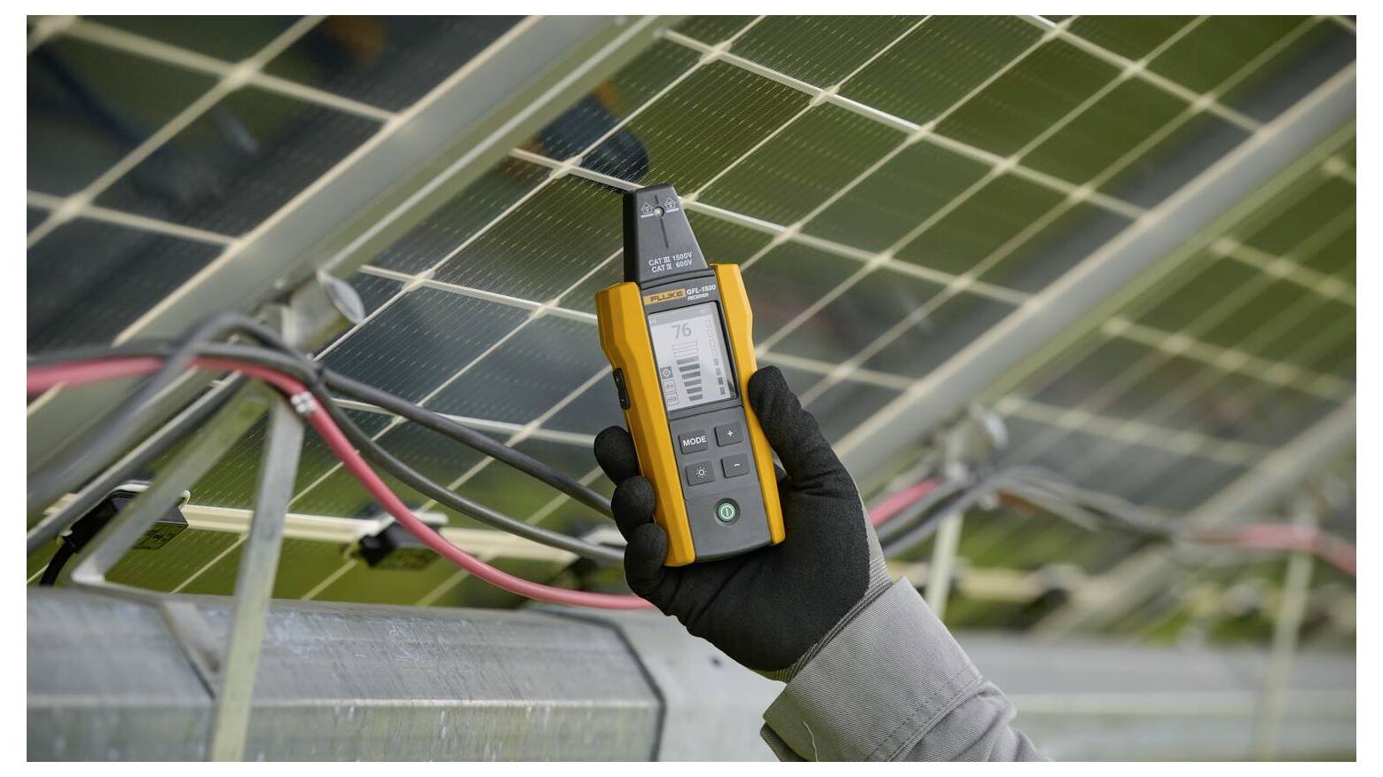 Eine Hand, die ein orange-schwarzes Gerät hält, das die Ausgabe eines Solarpanels misst. Das Gerät zeigt digitale Messungen an.