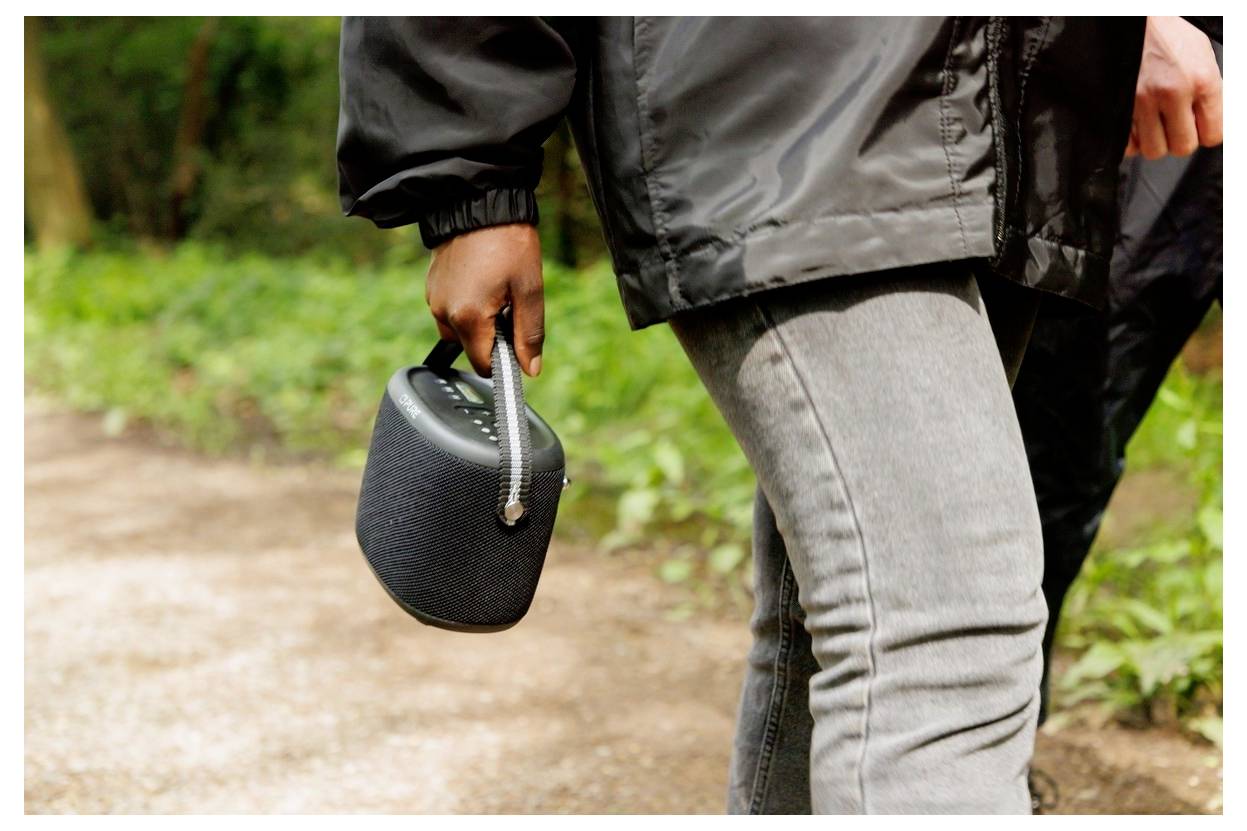 Eine Person, die im Freien geht und einen kleinen schwarzen tragbaren Lautsprecher an seinem Trageriemen hält, mit Grünpflanzen im Hintergrund.