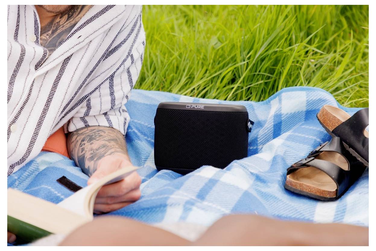 Eine Person in einem gestreiften Shirt liest ein Buch auf einer blauen Picknickdecke, neben der ein schwarzer Lautsprecher steht, in einem grasigen Bereich.
