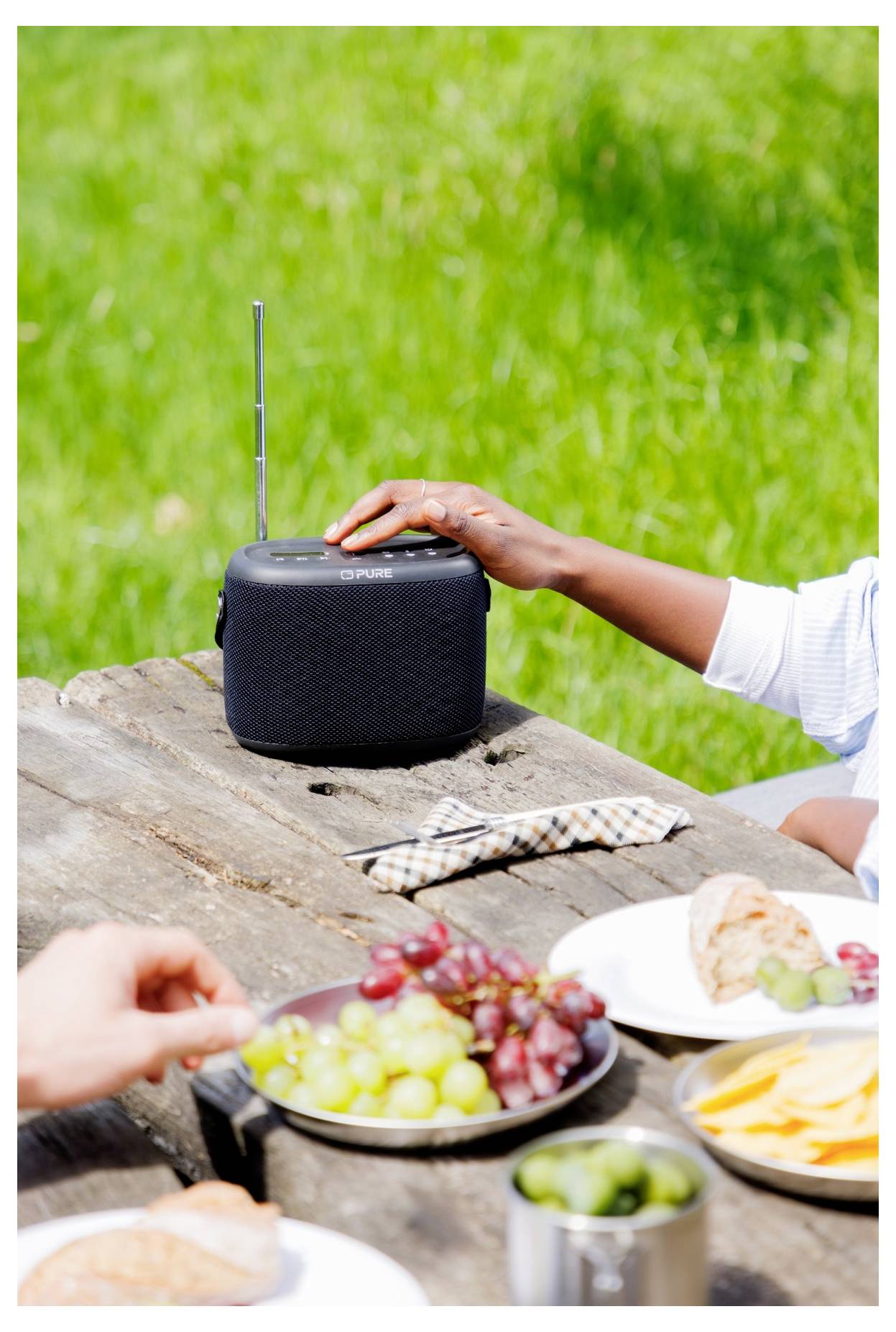 Eine Person justiert ein tragbares Radio auf einem Picknicktisch, der mit Tellern voller Trauben, Käse und Brot vor einem grünen Rasengrund bestückt ist.