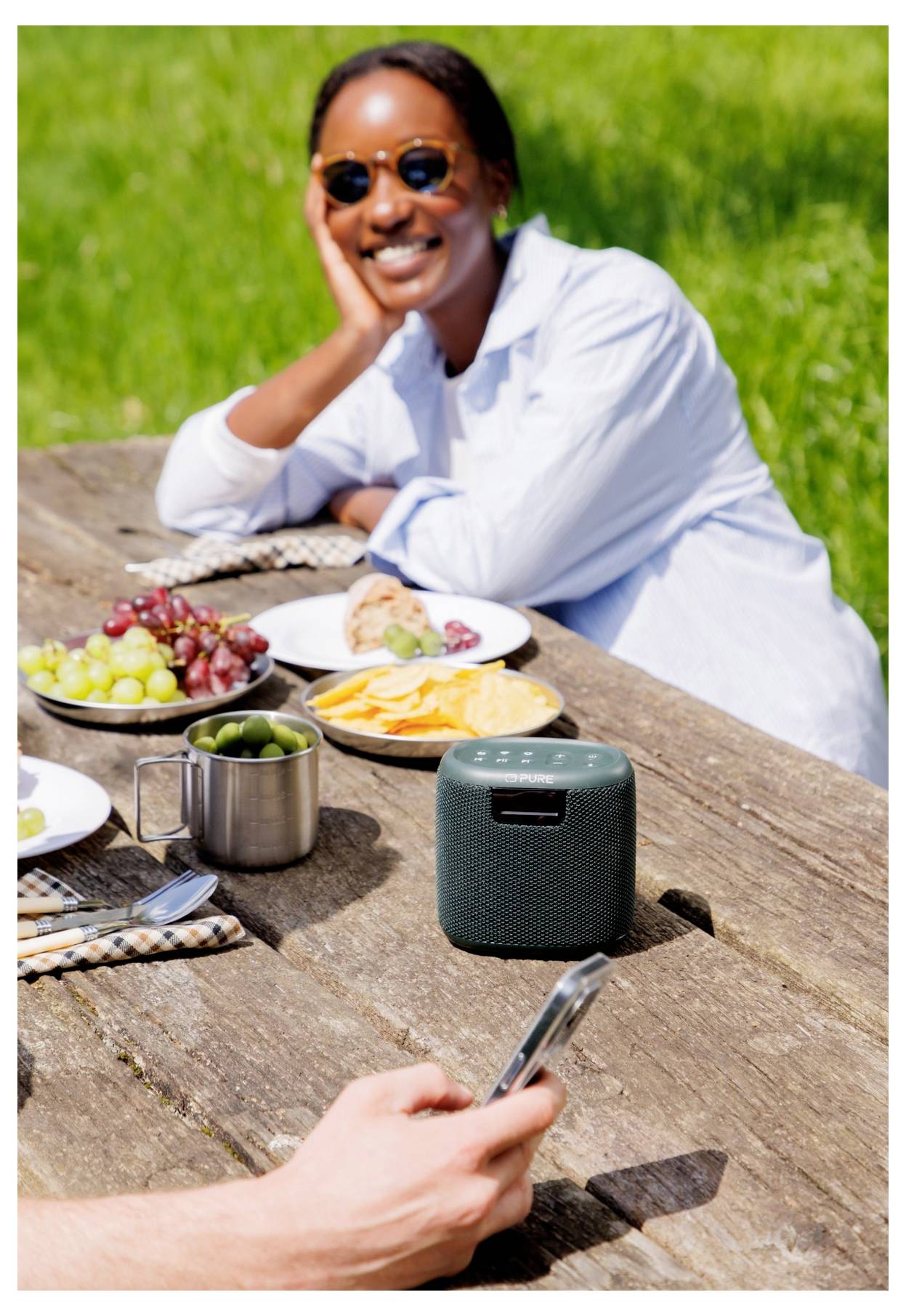 Eine Person mit Sonnenbrille lächelt an einem Picknicktisch im Freien, mit Essen, einem Lautsprecher und einer weiteren Hand, die ein Telefon hält, im Bild.
