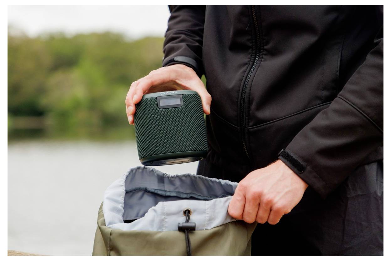 Eine Person in einer dunklen Jacke stellt einen tragbaren Lautsprecher in einen Rucksack im Freien, in der Nähe eines Gewässers mit Bäumen im Hintergrund.