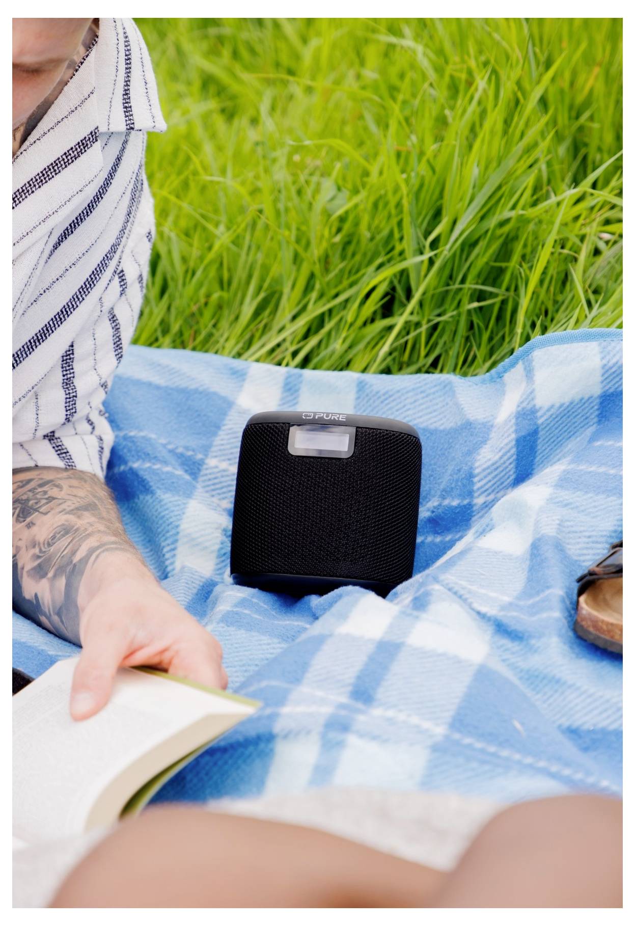 Eine Person liegt auf einer blau karierten Decke und liest ein Buch neben einem kleinen schwarzen Lautsprecher auf Gras.