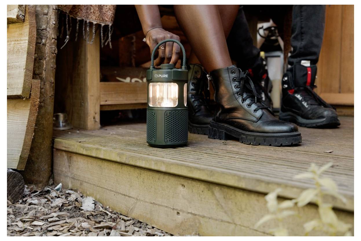 Eine Person in schwarzen Stiefeln sitzt auf einer Holzterrasse und hält eine grüne Laterne, was auf eine Outdoor- oder Campingaktivität hindeutet.