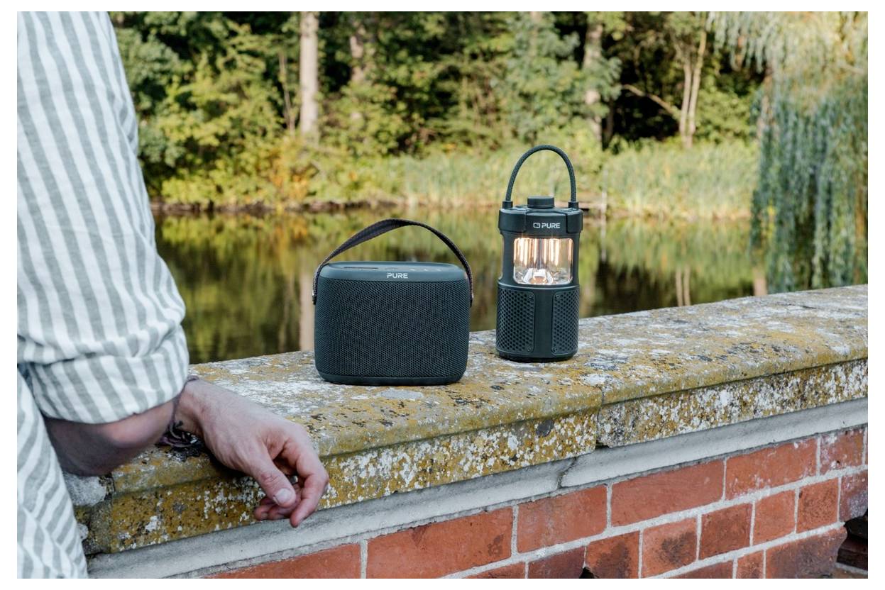 Eine Person in einem gestreiften Hemd steht neben einer Ziegelwand, auf der ein tragbarer Lautsprecher und eine Laterne stehen, in einer bewaldeten Seelandschaft.