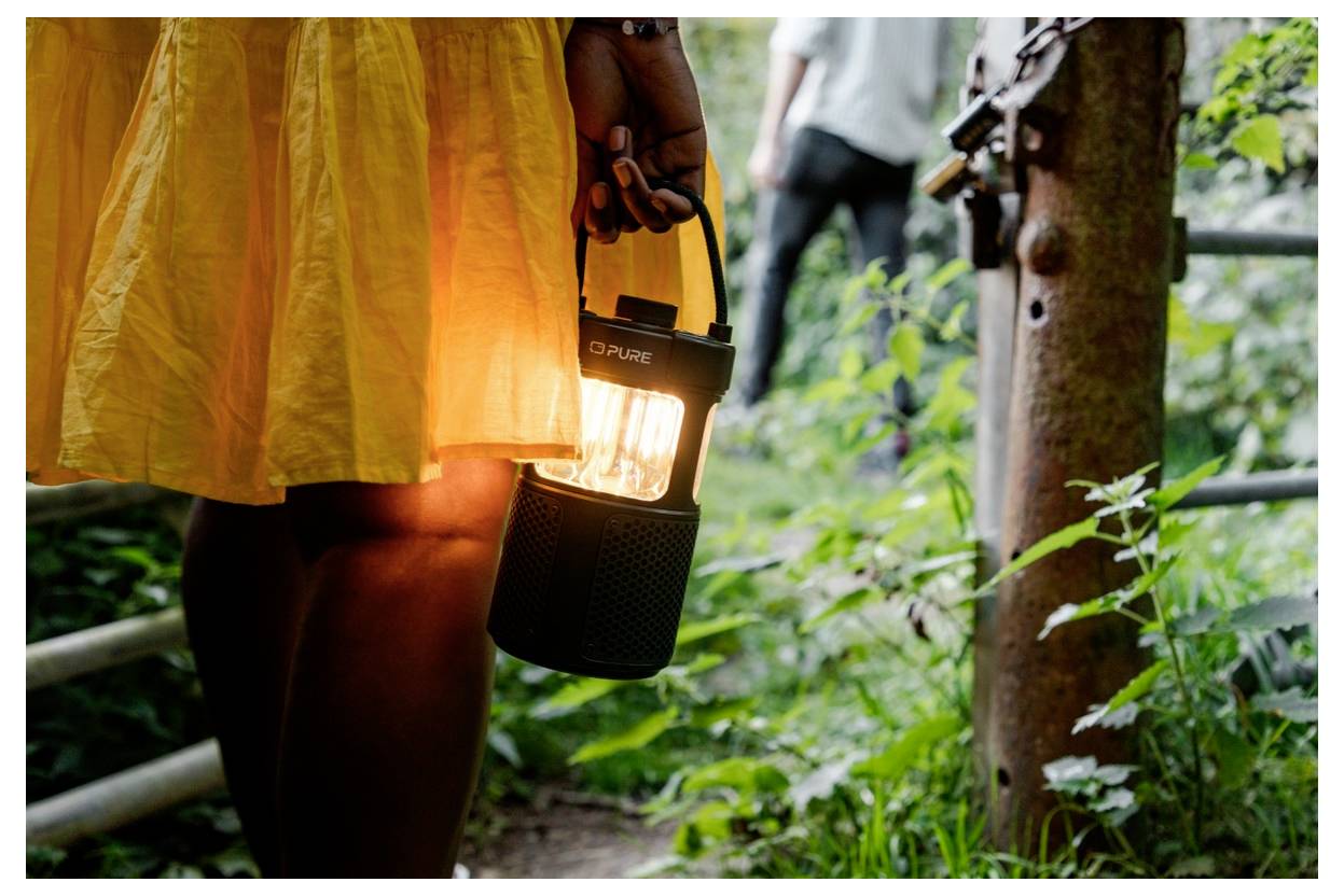 Eine Person in einem gelben Kleid hält eine brennende Laterne, während sie einen üppig grünen Weg entlanggeht, mit einer weiteren Person im Hintergrund sichtbar.