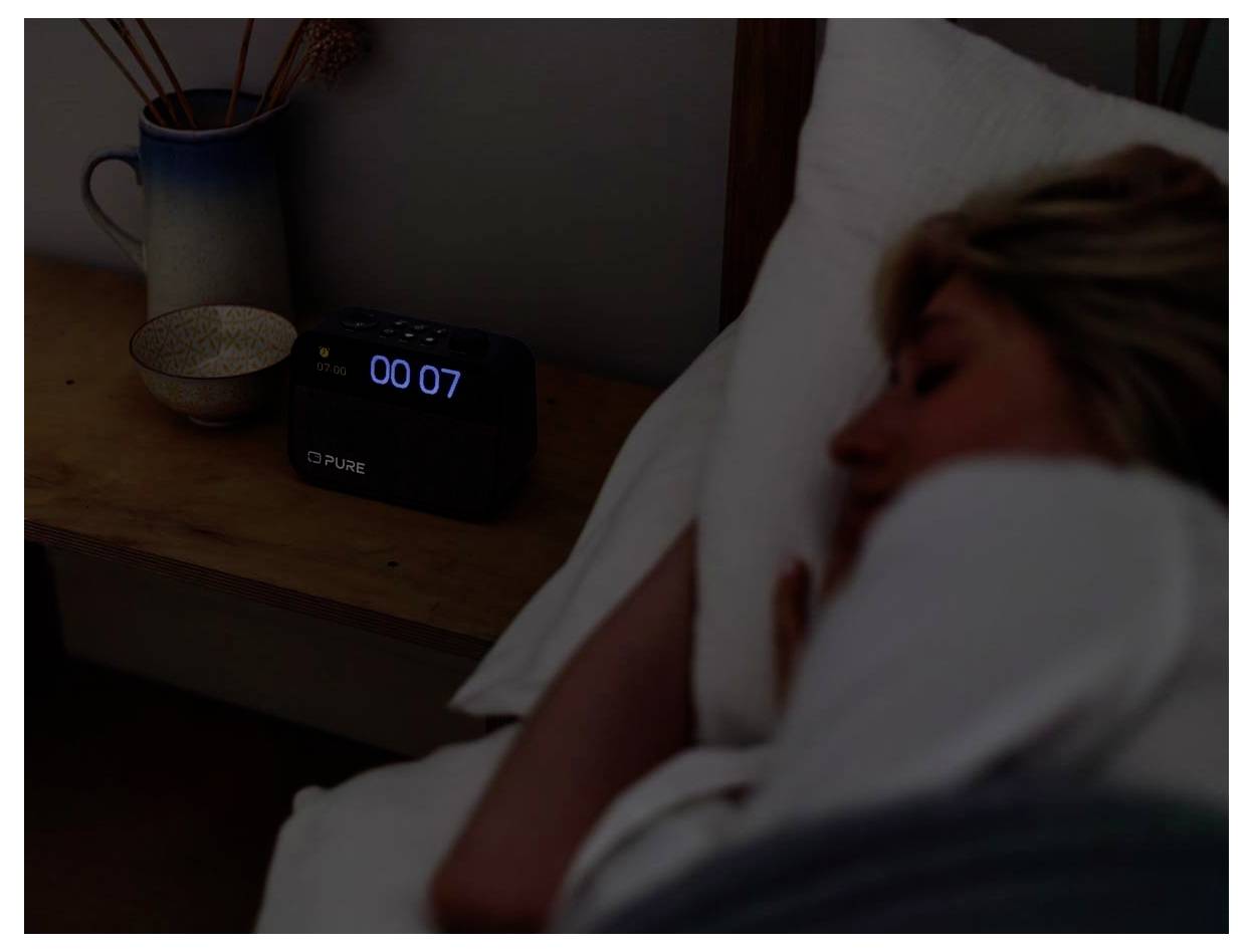 Eine Person schläft im Bett, während ein Wecker auf dem Nachttisch '00:07' anzeigt. Eine Tasse und eine Vase befinden sich ebenfalls auf dem Tisch.