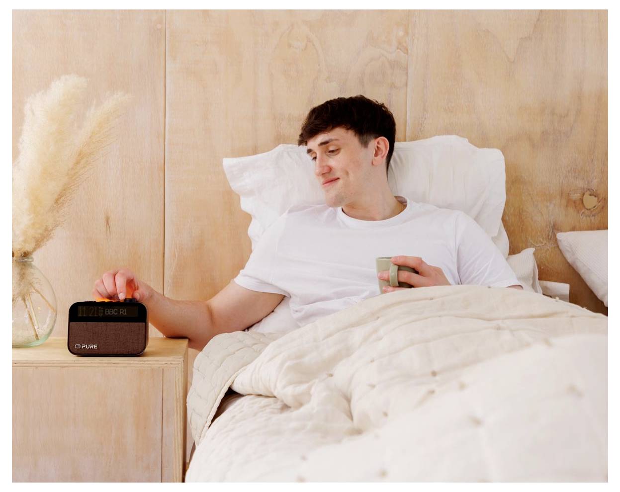 Eine Person im Bett, die lächelt, während sie einen Wecker auf dem Nachttisch ausschaltet. Sie hält eine Tasse und ist von gedeckt-farbener Bettwäsche umgeben.