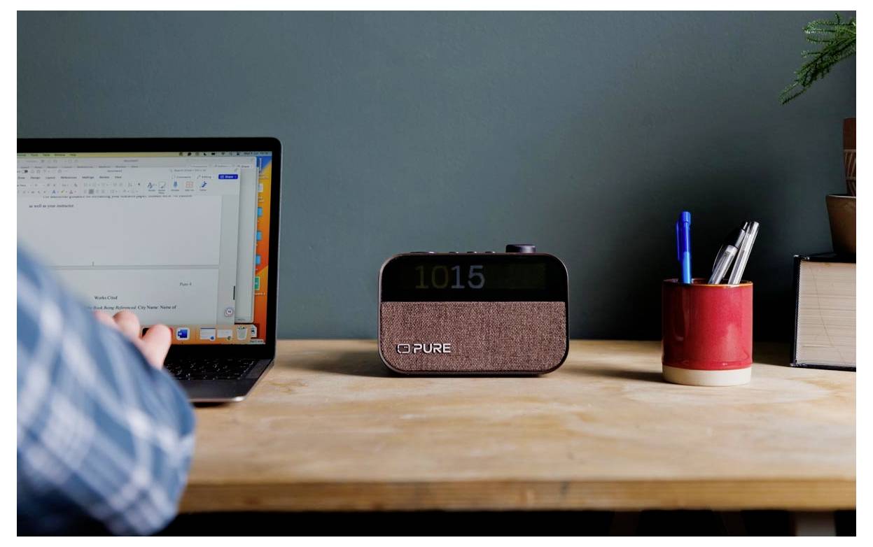 Eine Person tippt an einem Laptop auf einem Holzschreibtisch, neben einer digitalen Uhr, die '10:15' anzeigt, und einem roten Stifthalter, der Stifte und eine Pflanze enthält.