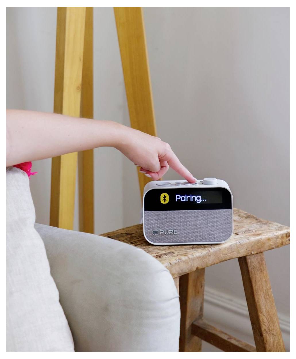 Eine Hand drückt einen Knopf auf einem kleinen Lautsprecher, auf dessen Bildschirm "Verbinden..." angezeigt wird, was einen Bluetooth-Verbindungsversuch signalisiert.