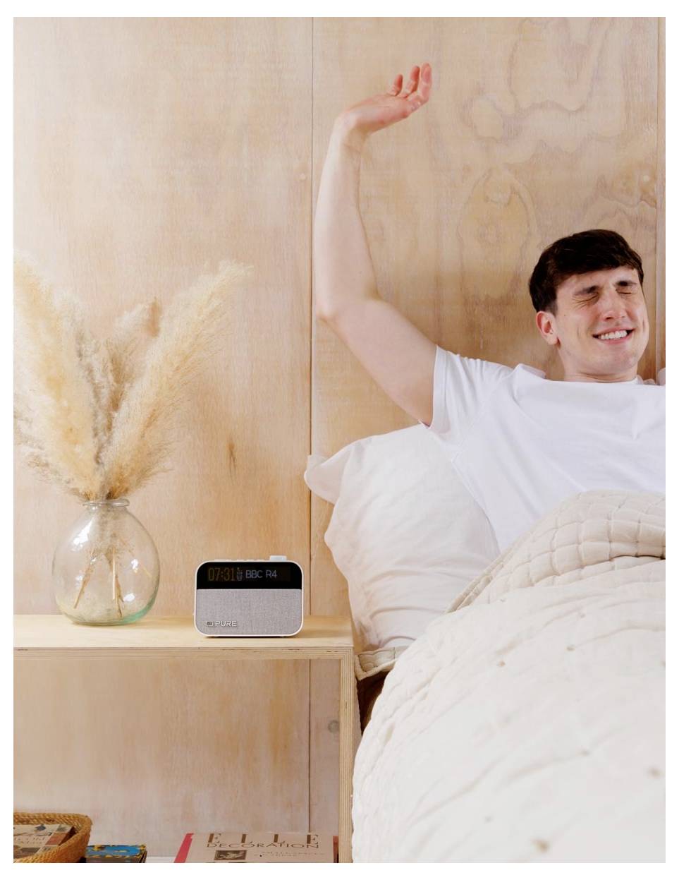 Eine Person streckt sich und lächelt im Bett und deutet auf einen angenehmen Morgen hin. Auf dem Nachttisch steht eine Vase mit getrockneten Blumen und ein digitales Radio.