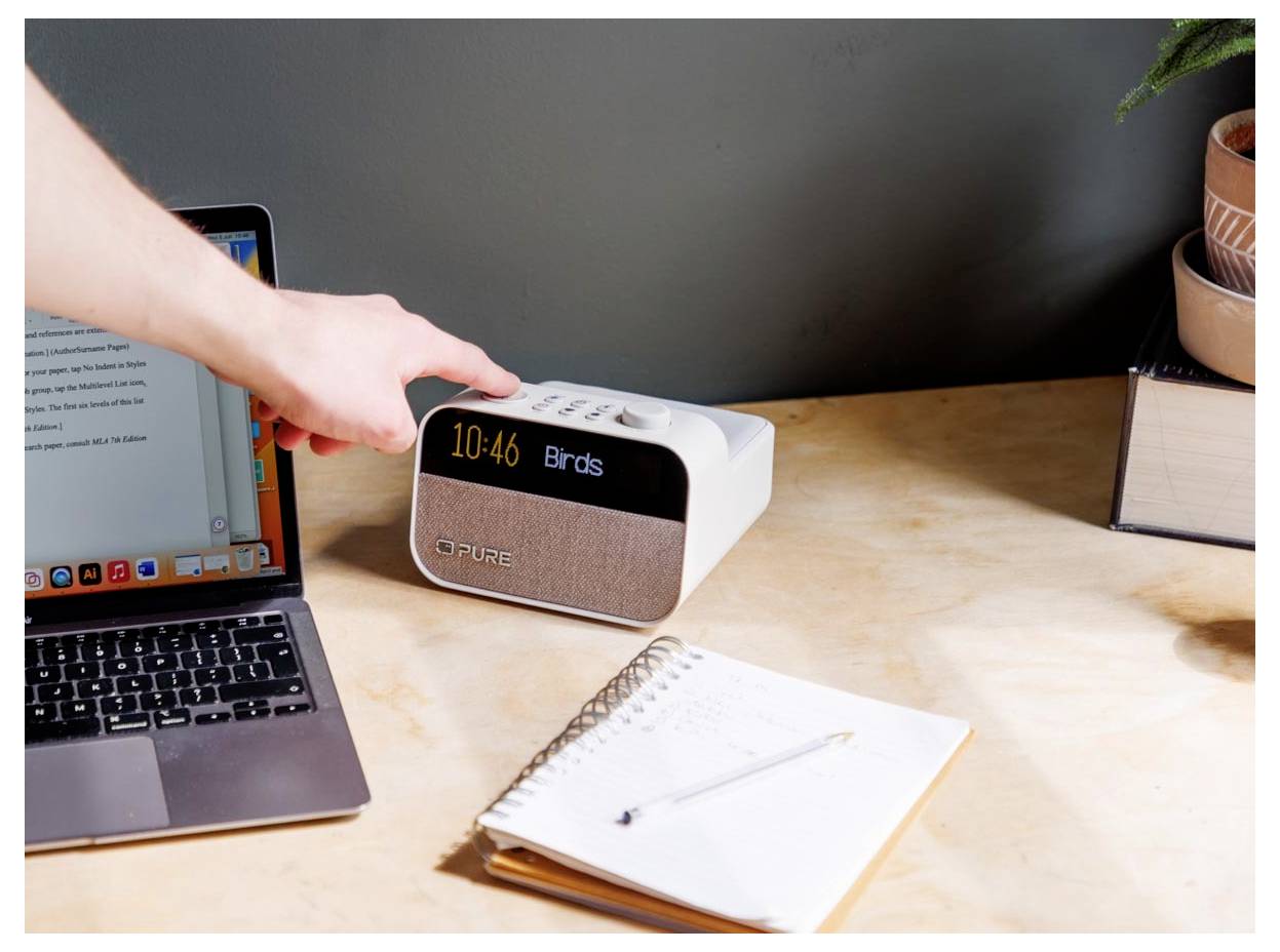 Eine Hand zeigt auf eine digitale Uhr, die '10:46' und 'Birds' anzeigt, neben einem Laptop und einem Notizbuch mit einem Stift auf einem Holzschreibtisch.