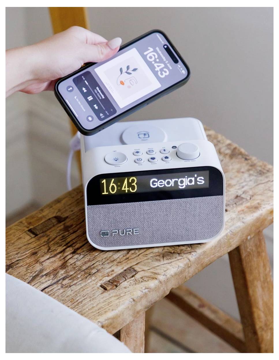 Eine Person hält ein Telefon mit einer Musik-App über ein digitales Radiowecker. Die Uhr zeigt '16:43' und den Namen 'Georgia's'.