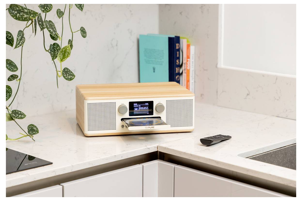 Ein hölzernes digitales Radio mit einem kleinen Display und einem nach vorn gerichteten Lautsprecher steht auf einer Küchenarbeitsplatte neben einer Fernbedienung und einer Pflanze.