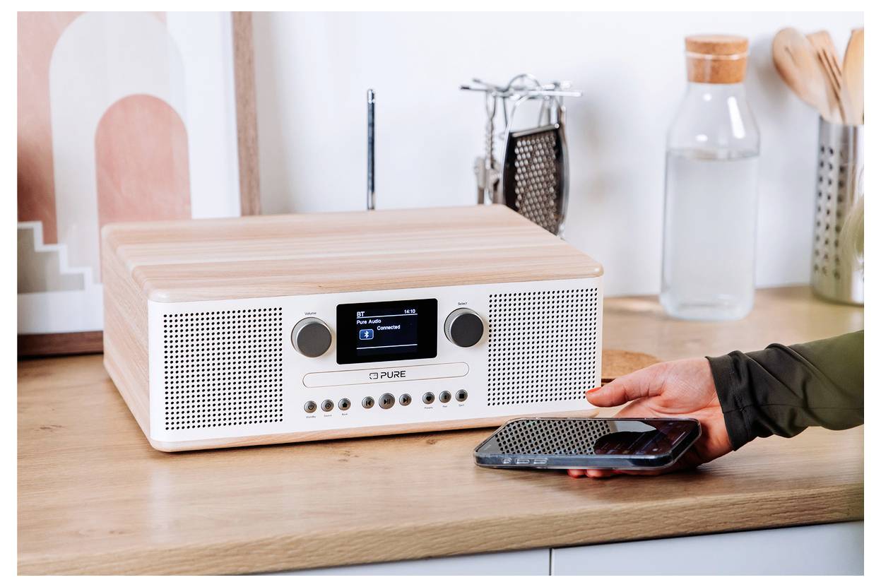 Eine moderne Holzradio auf einer Küchenarbeitsfläche mit einer Menschenhand, die mit einem Smartphone interagiert und drahtlose Konnektivität andeutet.